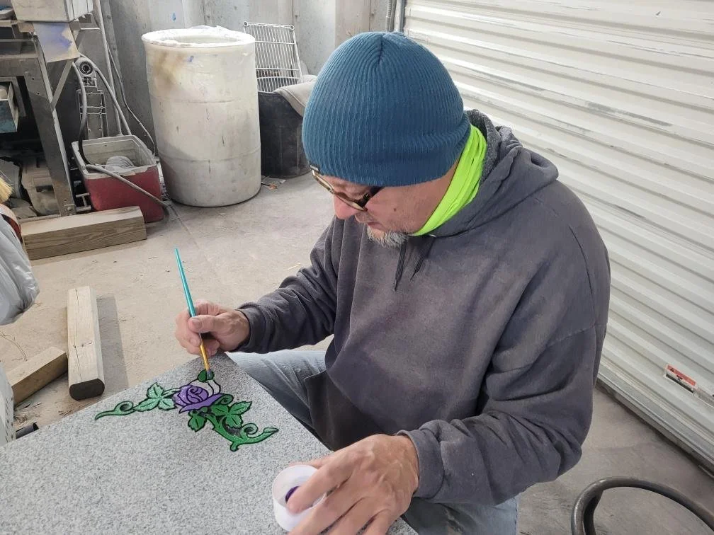 A man wearing a blue beanie, glasses, a gray hoodie, and a neon green shirt is painting a decorative design of purple roses and green leaves on a gray stone or concrete surface inside a workshop or garage.