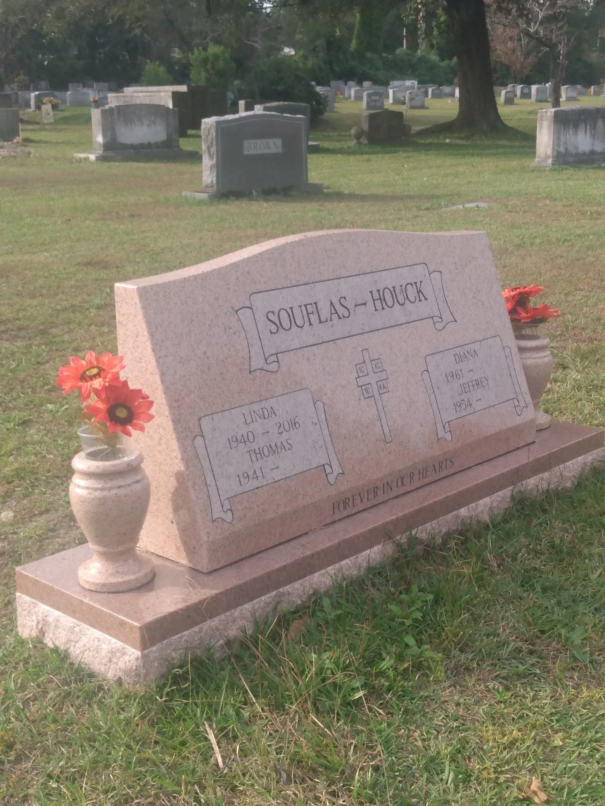 Gravestone in a cemetery with flowers on each side and a large tree in the background.