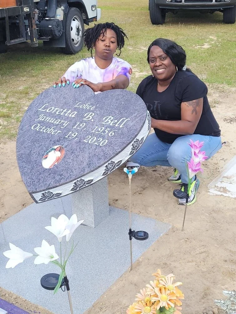 Two women kneeling beside a heart-shaped grave marker with flowers and small memorial items, at a cemetery.