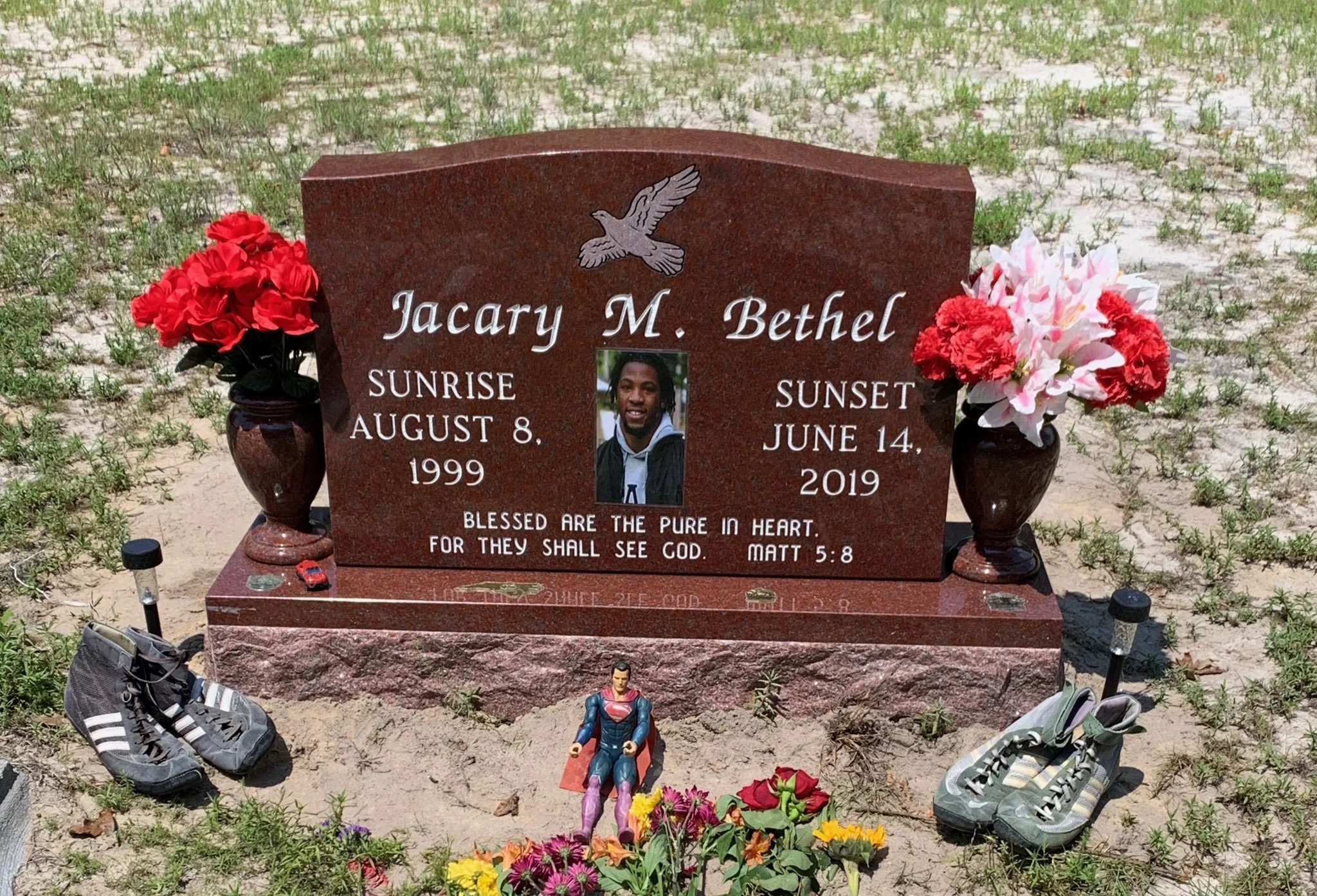 A gravesite with a red granite headstone for Jacary M. Bethel, who was born on August 8, 1999, and died on June 14, 2019. The headstone features an engraved dove and a photograph of Jacary. There are flowers and toy figurines, including Superman, pla