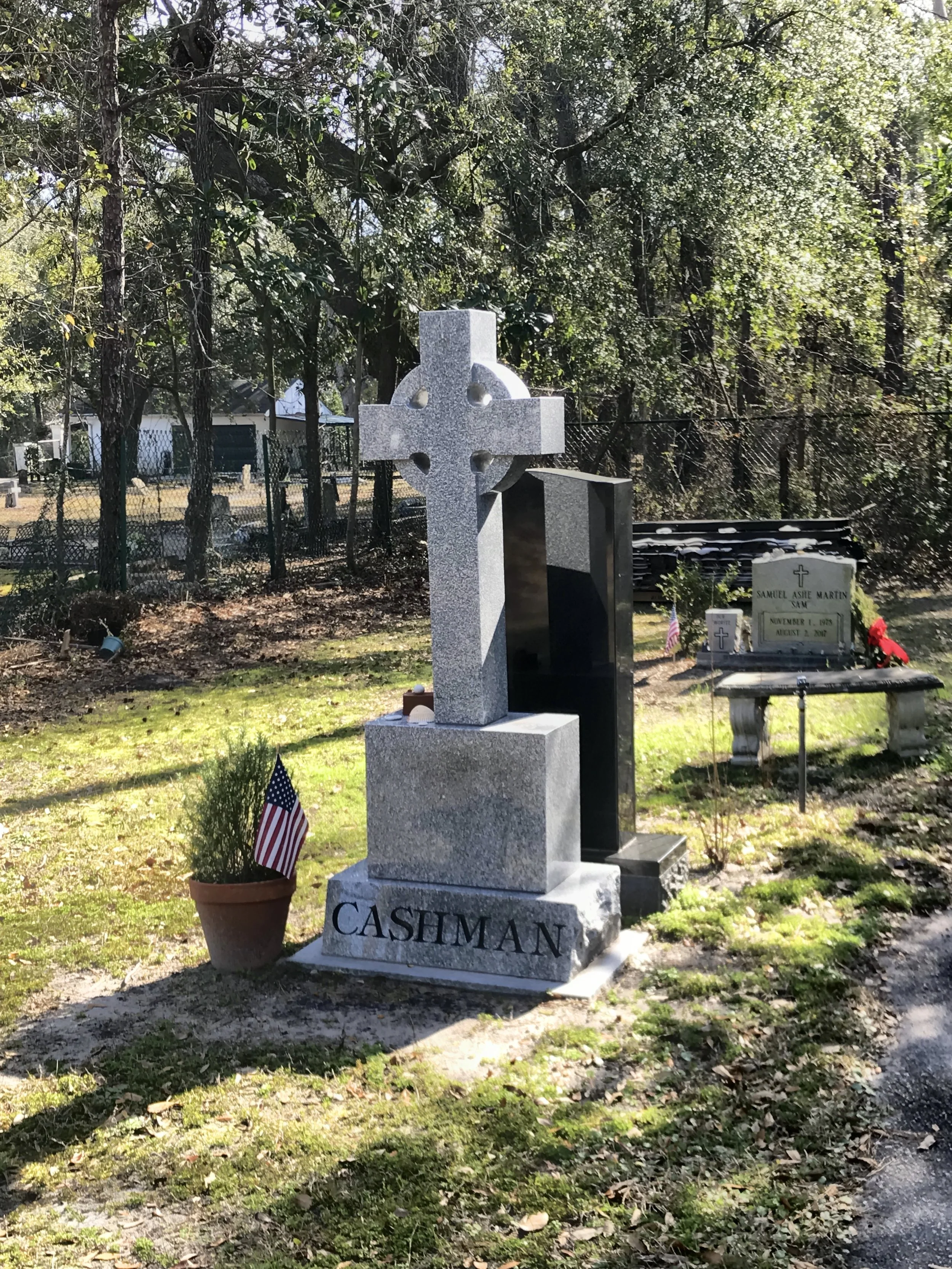 A cemetery with gravestones and a stone Celtic cross monument inscribed with the name 'Cashman'. A small American flag and a potted plant are placed in front of the monument. Trees and other gravestones are visible in the background.