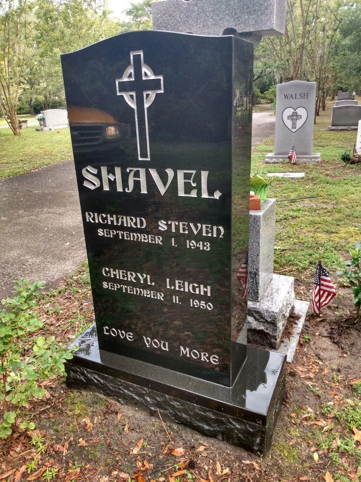 Black granite tombstone with a cross engraving, honoring Richard Steven Shavel (September 1, 1943) and Cheryl Leigh (September 11, 1950), with small American flags and a heartfelt message "Love You More" at the bottom. Located in a cemetery with othe