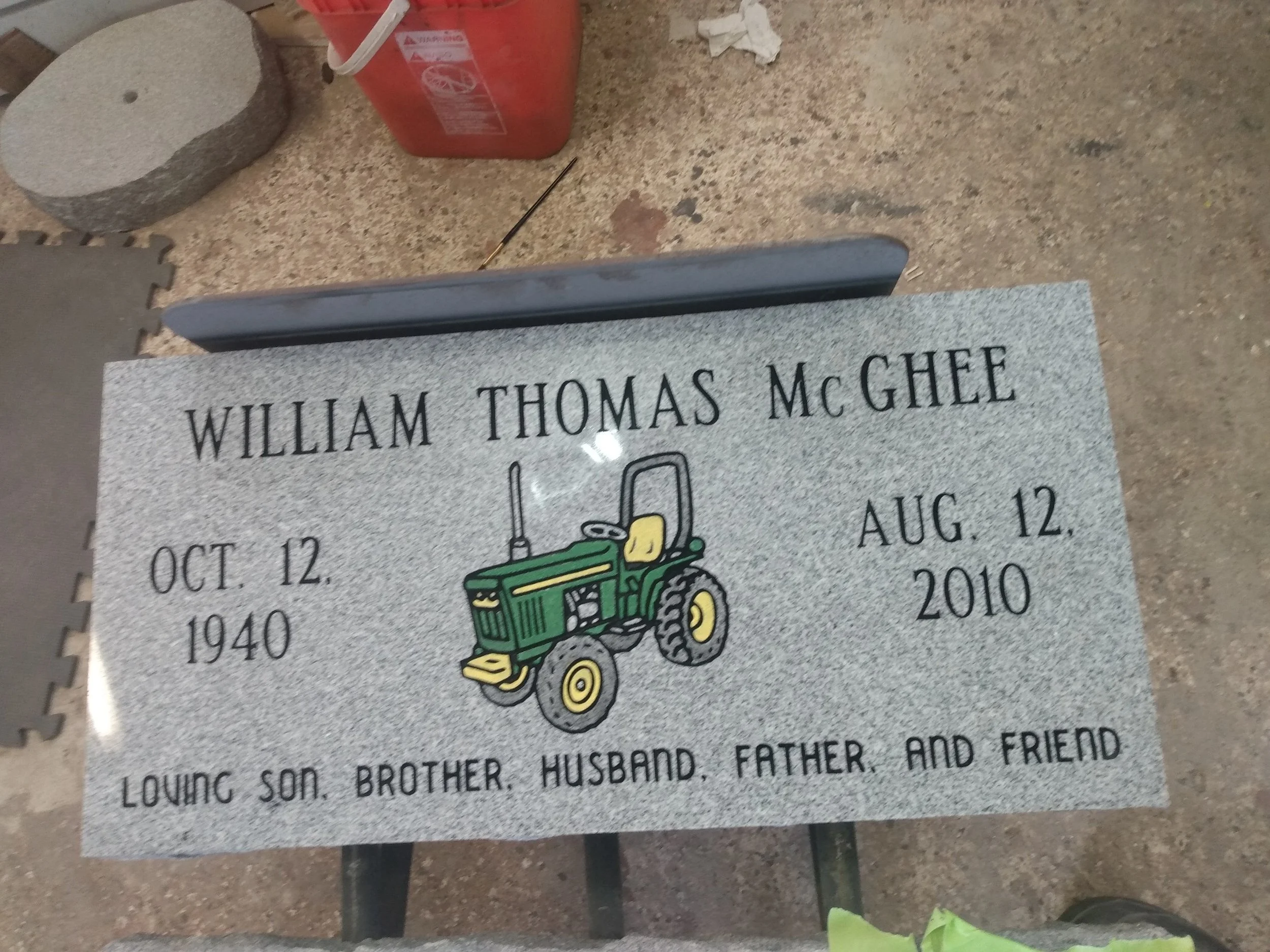 Gravestone with the name William Thomas McGhee, obituary dates October 12, 1940 to August 12, 2010, featuring a cartoon tractor illustration and a small stone placed nearby.