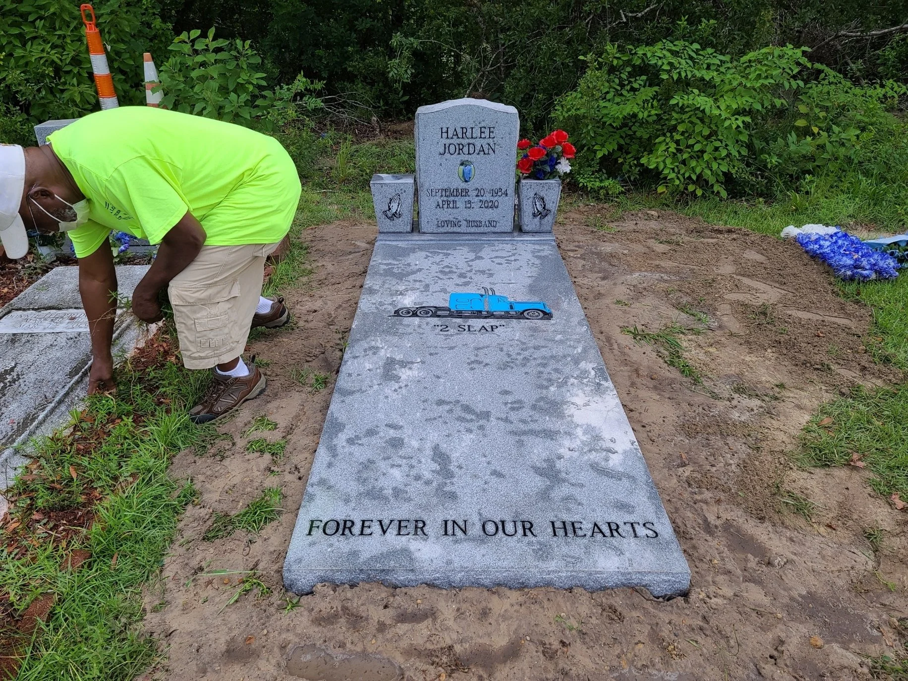 A man wearing a neon yellow shirt, beige shorts, a hat, and a face mask bending down next to a grave. The grave has a gray stone headstone inscribed with the name Harlee Jordan, birth and death dates, and a message "Loving Husband". The grave marker 