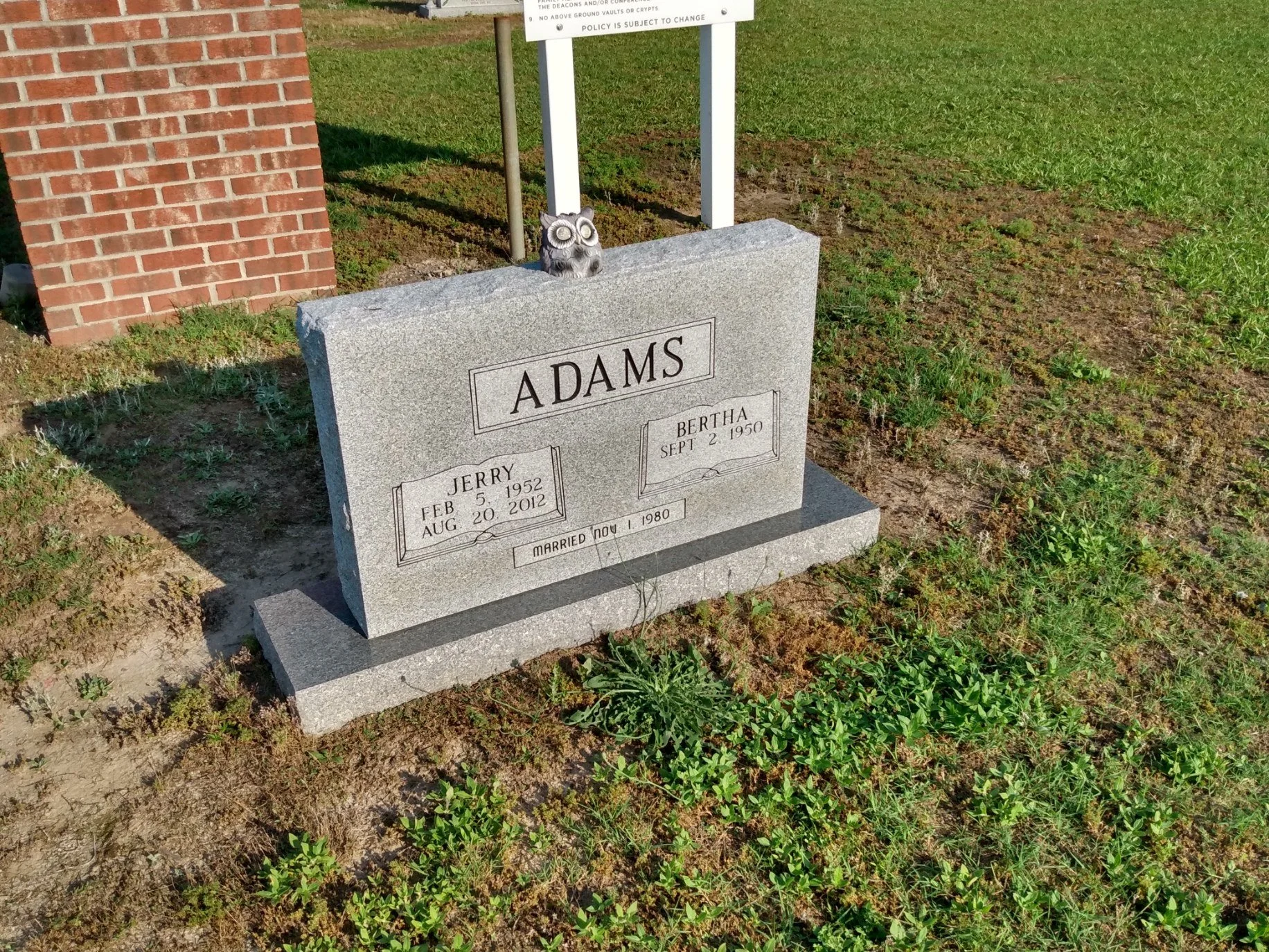 Gravestone with the surname 'Adams' engraved, indicating the names and birth and death dates of Jerry and Bertha Adams. The gravestone is situated in a grassy area with some green weeds and a brick structure nearby.