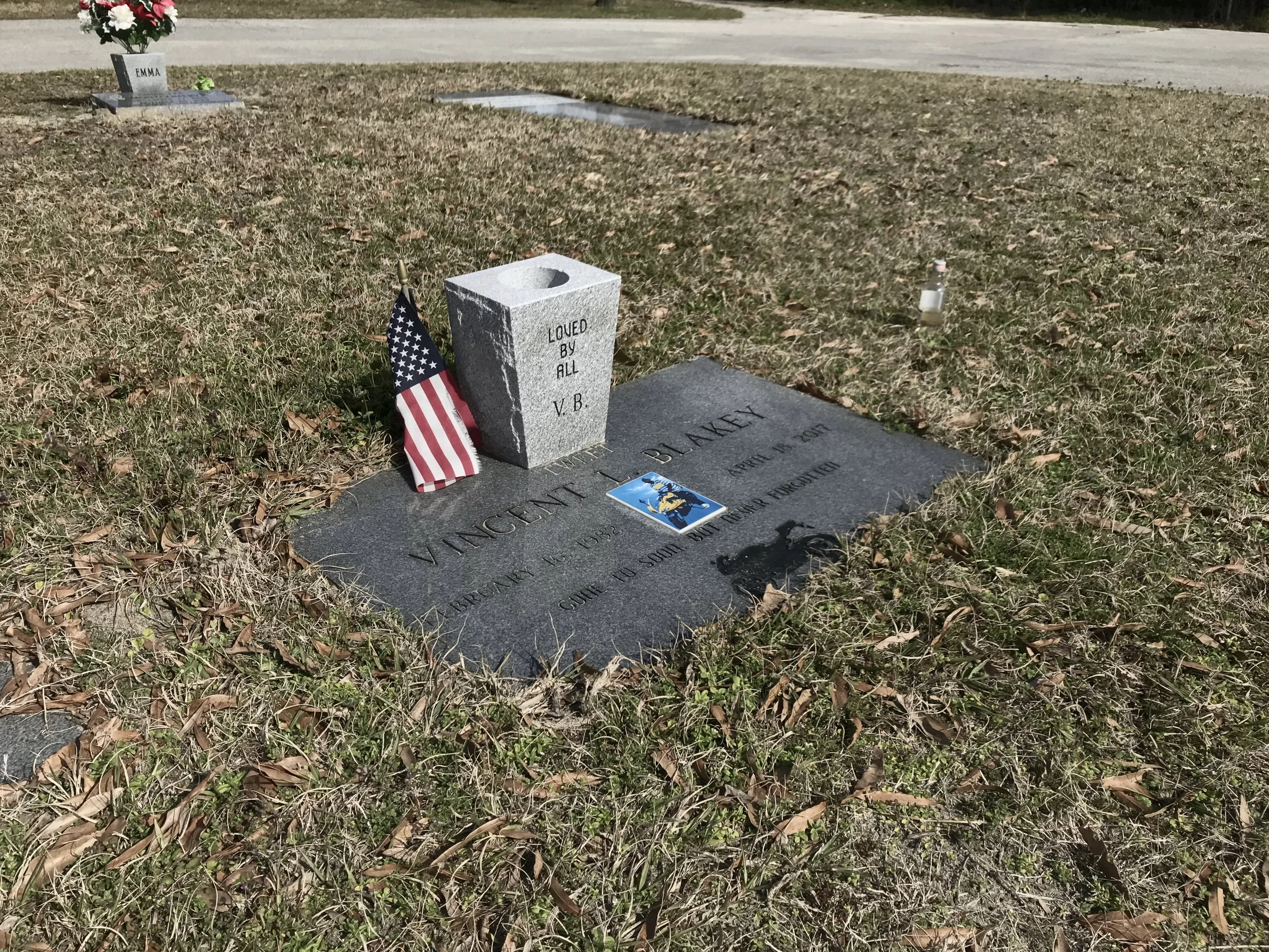 Gravestone in a cemetery with an American flag, a flower, and a small statue nearby. The grave marker is for Victor Blakeney, born April 16, 1932, and passed away on April 15, 2017.