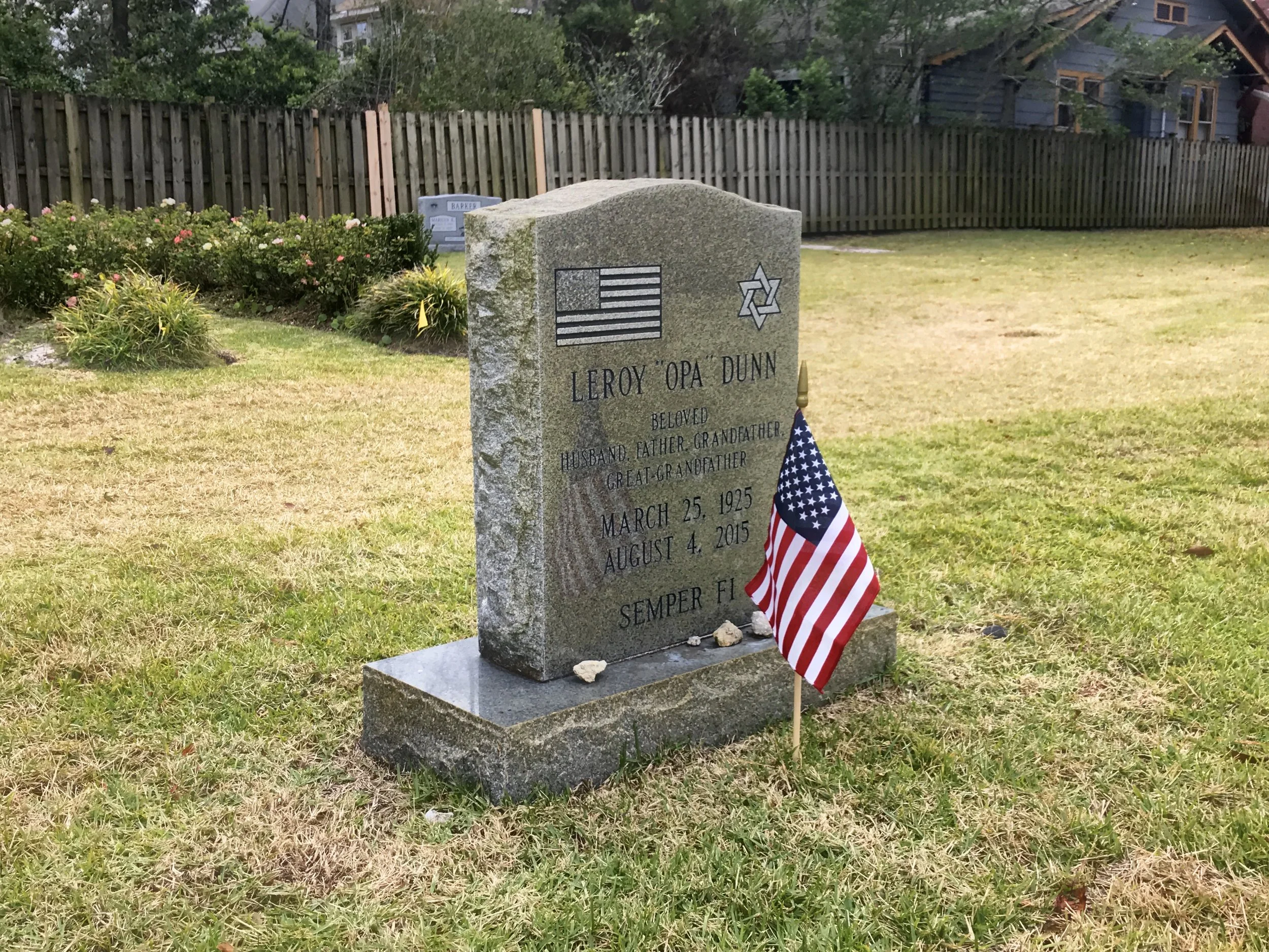 Gravestone in a cemetery with a US flag and small American flag decoration at the base, a flower bed with pink and white flowers, a wooden fence, and a house in the background.