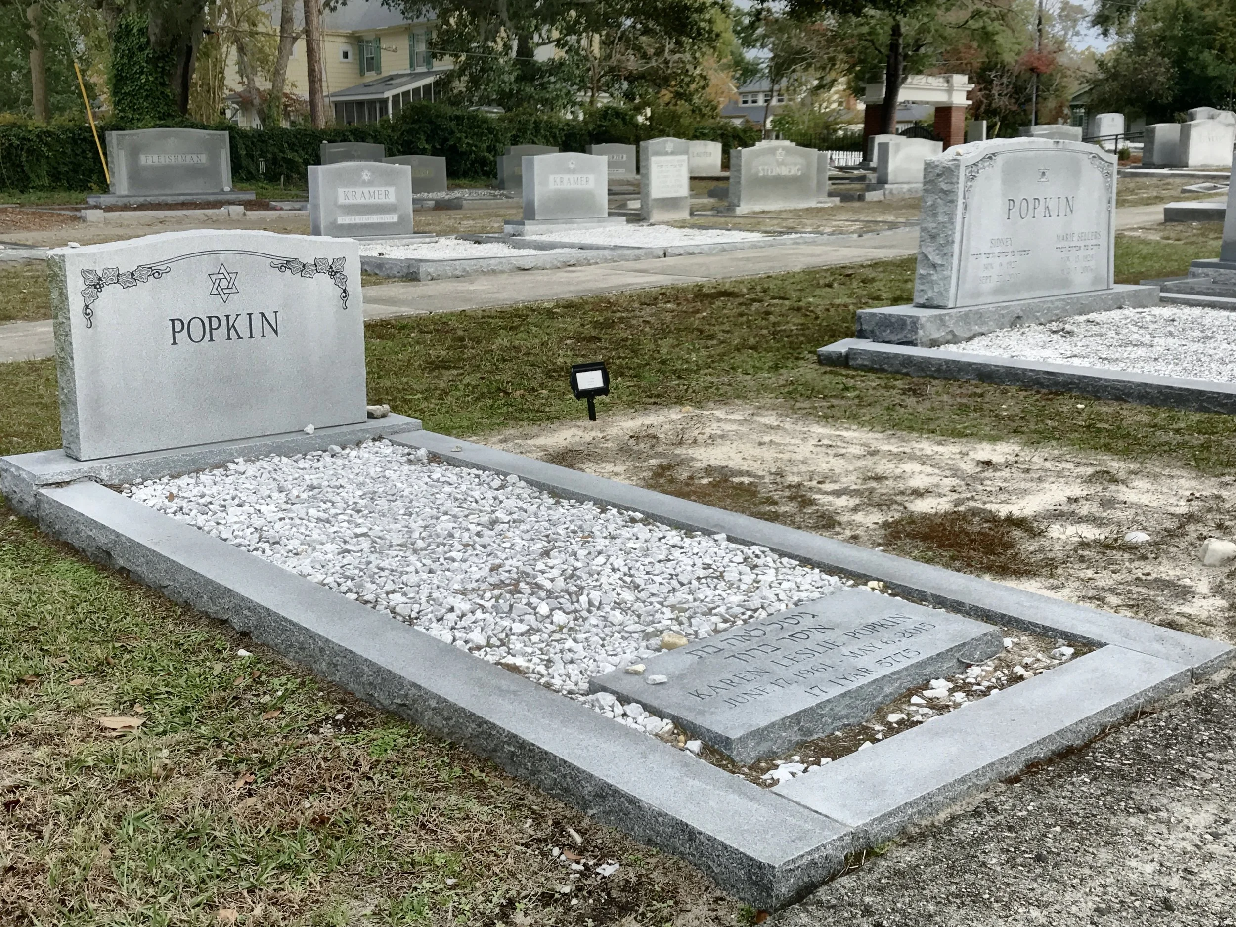 Multiple grave markers in a cemetery, including a gray headstone for Popkin with a Star of David symbol, surrounded by white gravel and grass. Other headstones are visible in the background, with some trees and houses beyond the cemetery grounds.