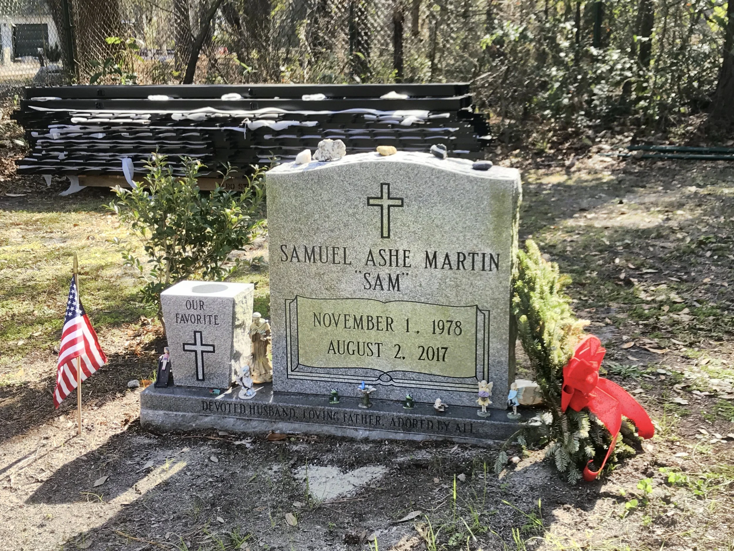 A grave with a gray headstone engraved with the name Samuel Ashe Martin, born November 1, 1978, died August 2, 2017. The grave is decorated with small toys, flags, flowers, and crosses. Behind the grave, there is a black structure with multiple level