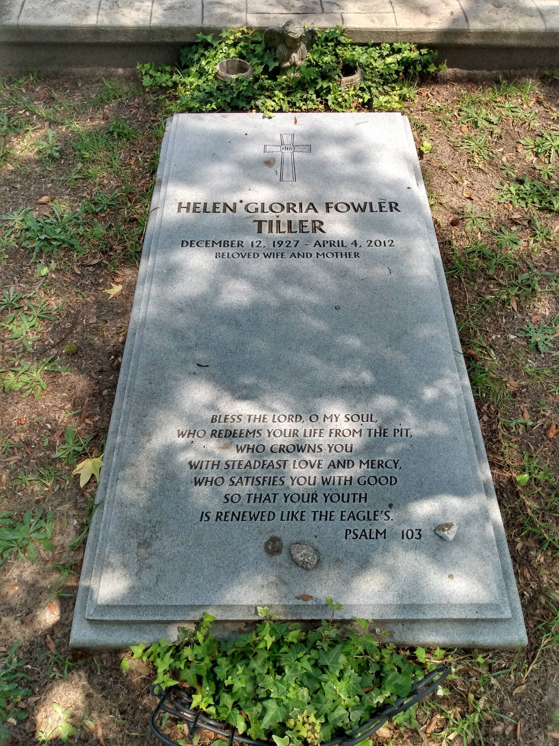 A gravestone for Helen Gloria Fowler Tiller, with an engraved cross and biblical verse from Psalm 103. The gravestone is surrounded by small plants and greenery.