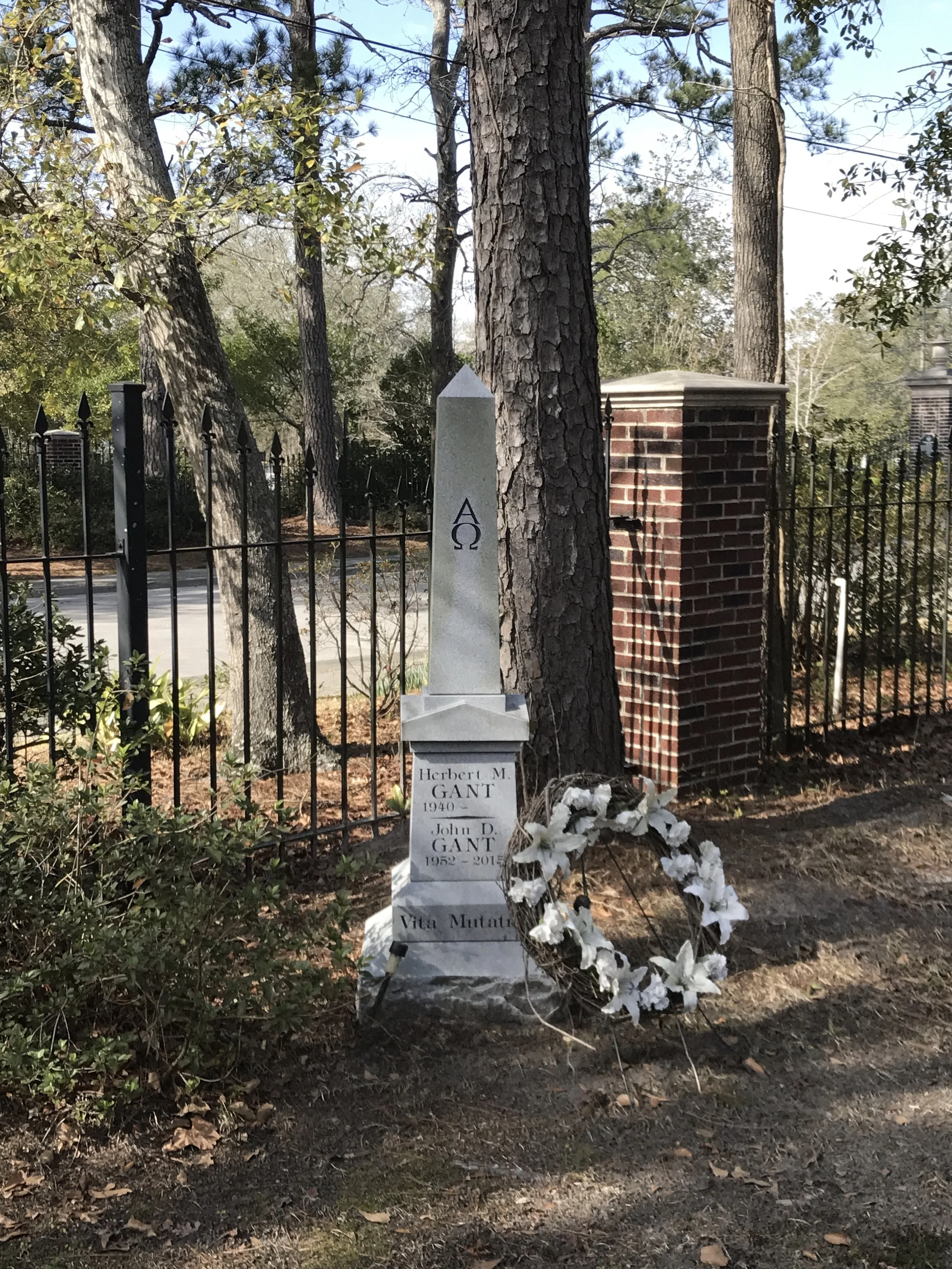 A cemetery grave marker engraved with the Omega symbol, dedicated to Herbert M. Gant (1940) and John D. Gant (1952-2015), with white floral wreath at the base, behind black metal fence and brick brick column, trees and sky in background.