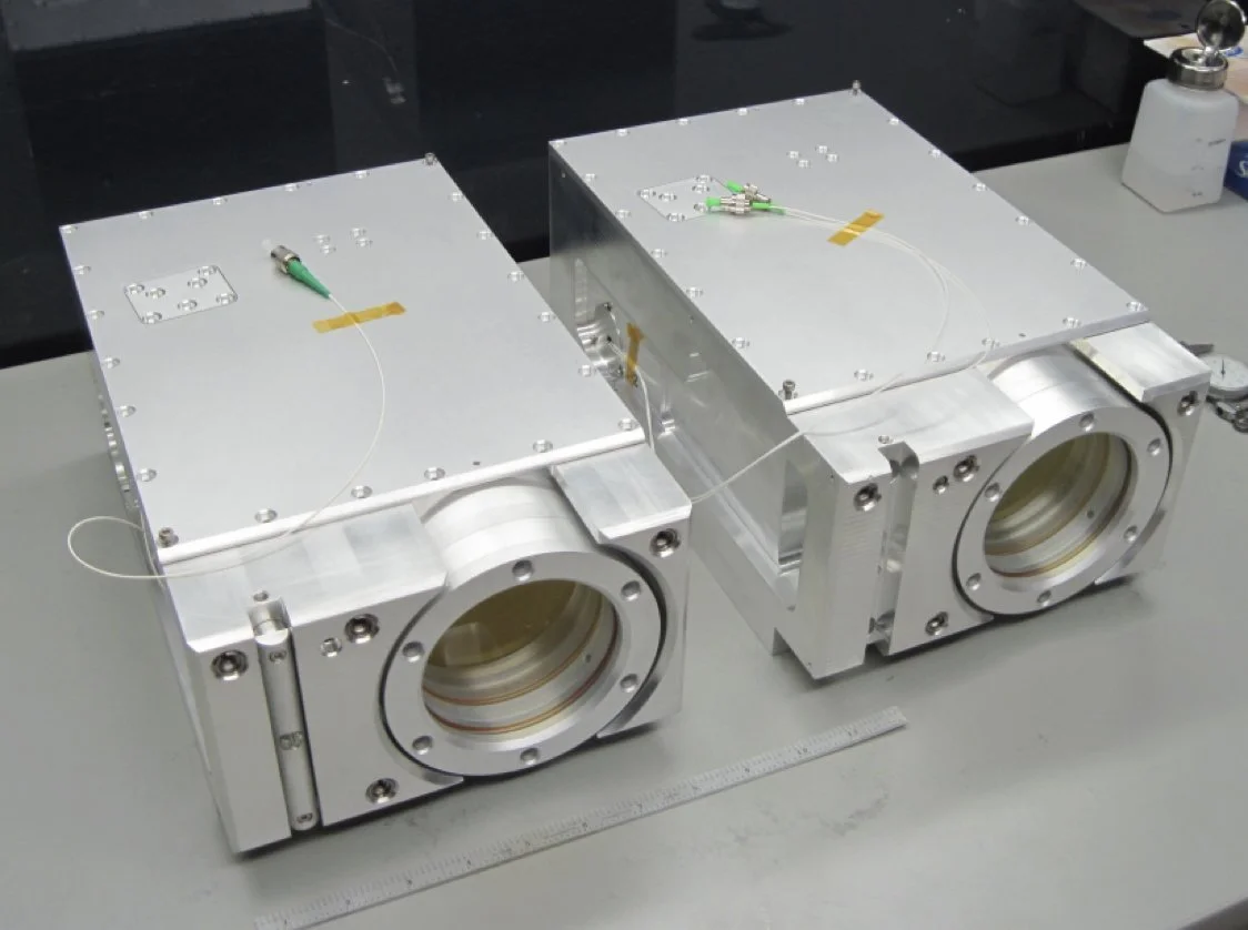 Two large metallic vacuum chambers with circular ports and attached sensors are positioned on a workbench in a laboratory.