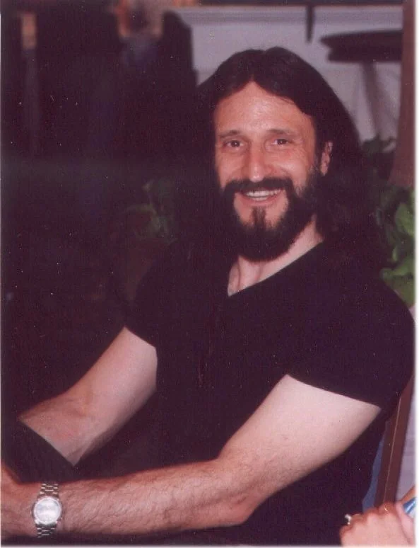 A man with long dark hair, a beard, and a mustache, smiling and wearing a black shirt, sitting at a table in an indoor setting.