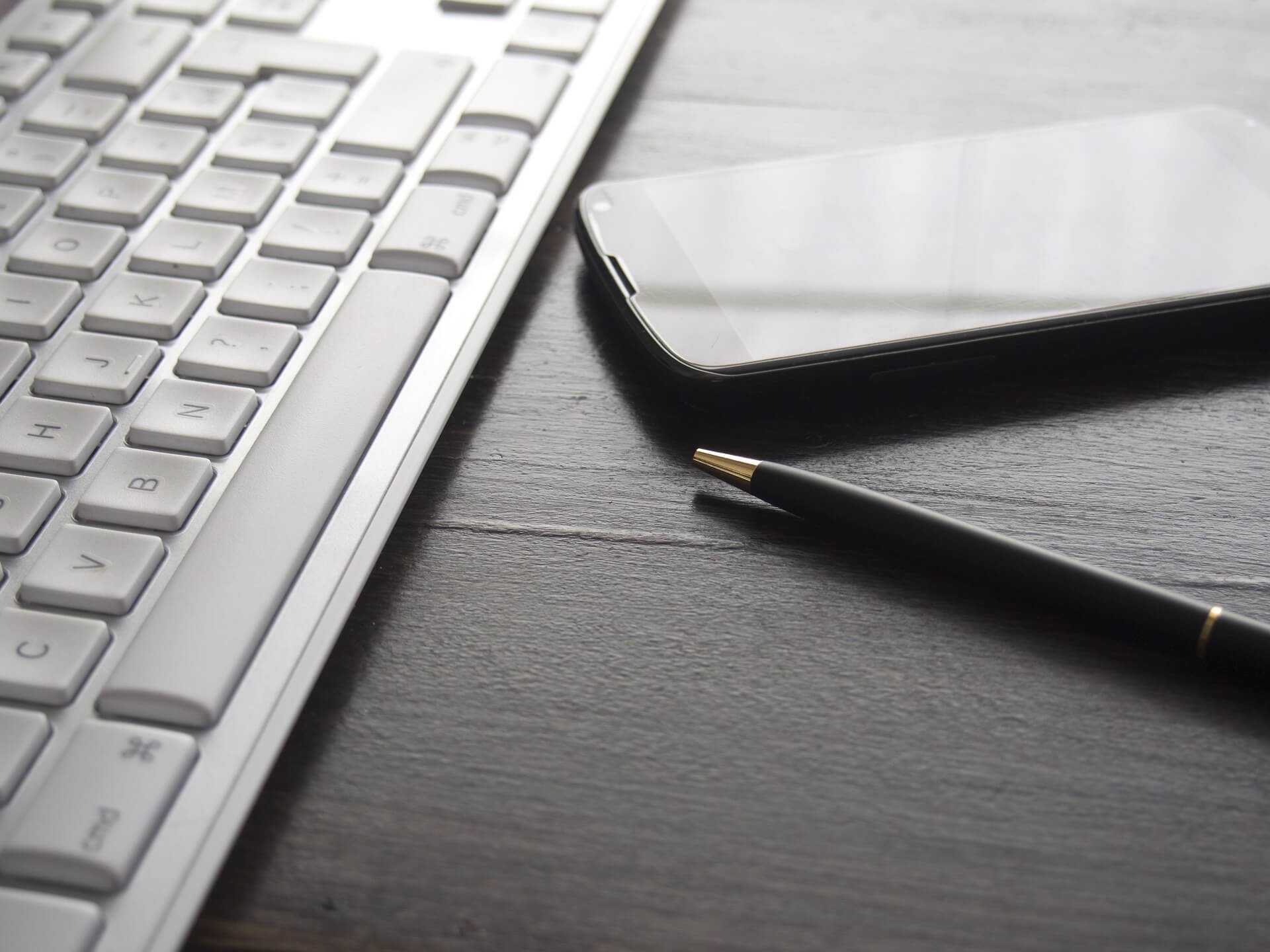 AppFolio Accounting | Close-up view of a white computer keyboard, a smartphone, and a black pen with gold tip on a dark wooden desk. | Northstar Trust Accounting | HOA Financials | Monthly Reconciliation | AppFolio | DRE Compliance