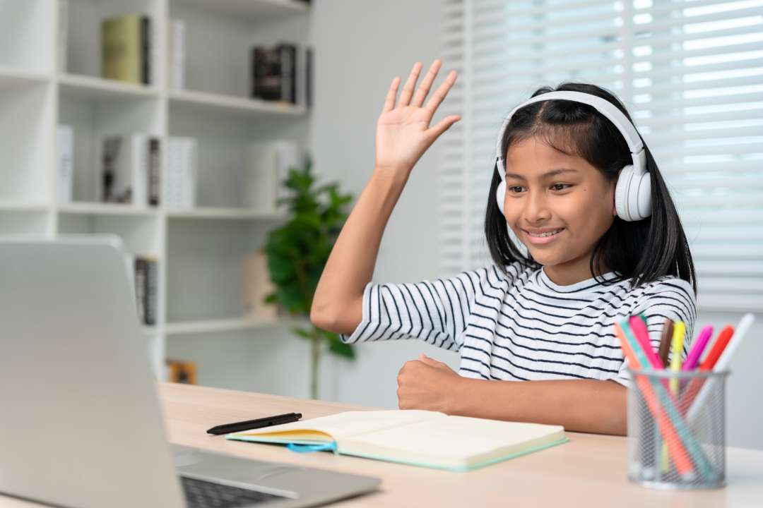 Young girl wearing headphones, smiling, raising hand, sitting at a desk with an open notebook, black pen, and a container of colorful pens, in a bright room with a laptop and bookshelves.