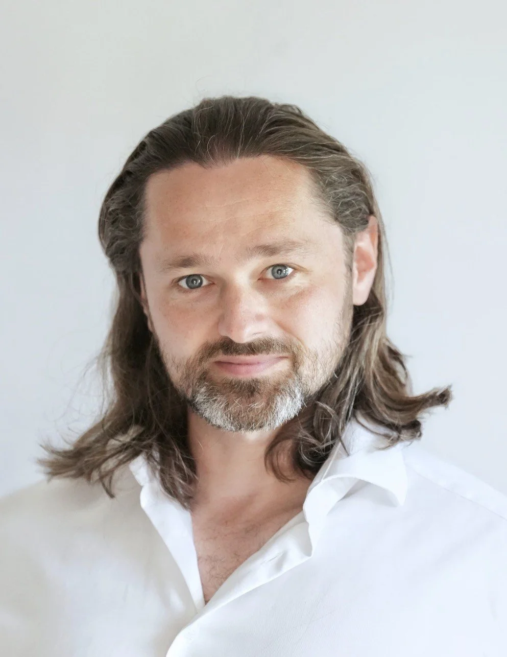 A man with long brown hair, a beard, and blue eyes, wearing a white shirt, posing against a plain light-colored background.