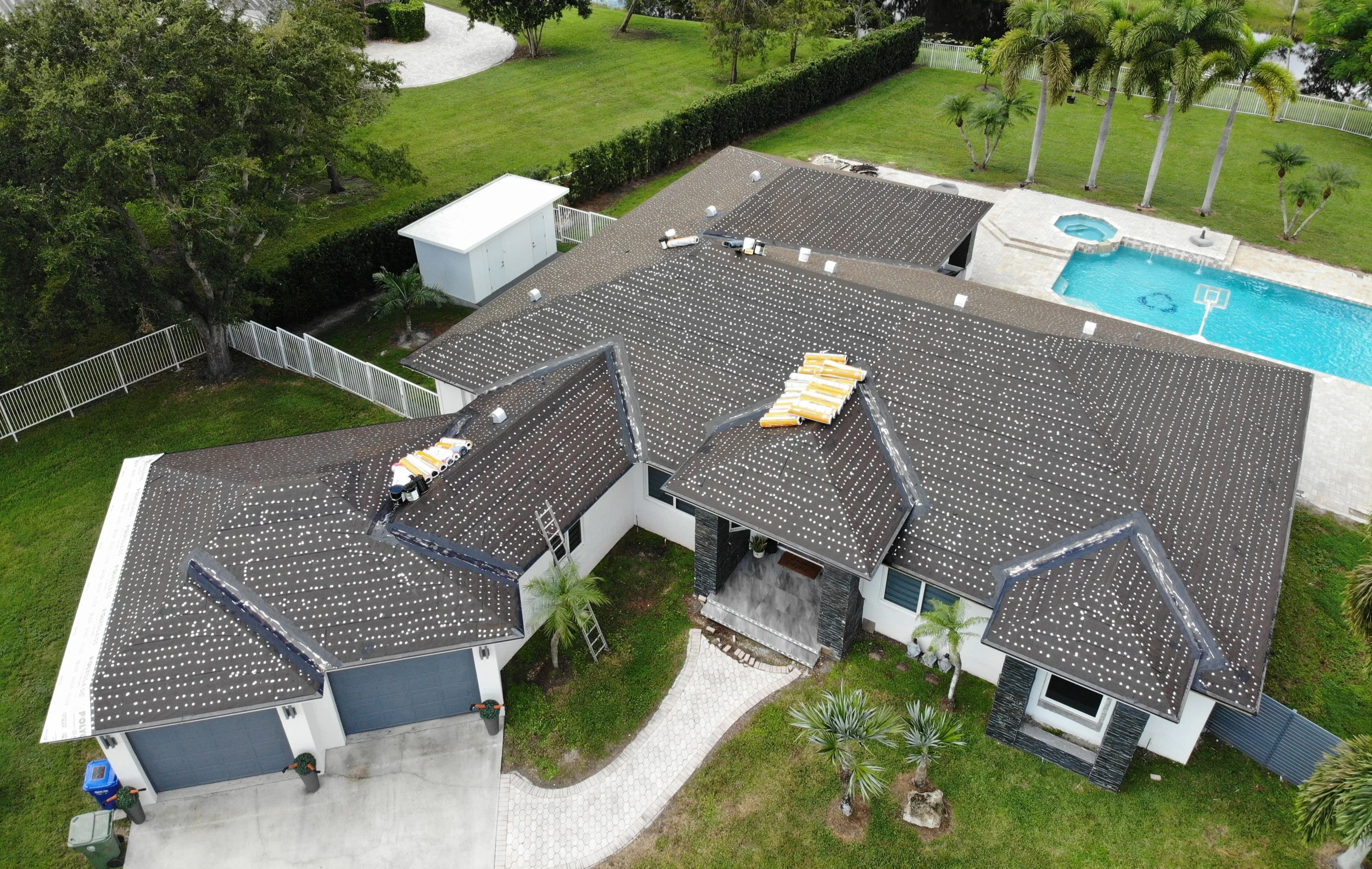 Aerial view of a house with a dark gray roof, a backyard with a swimming pool, hot tub, and various trees and greenery, surrounded by a white fence.