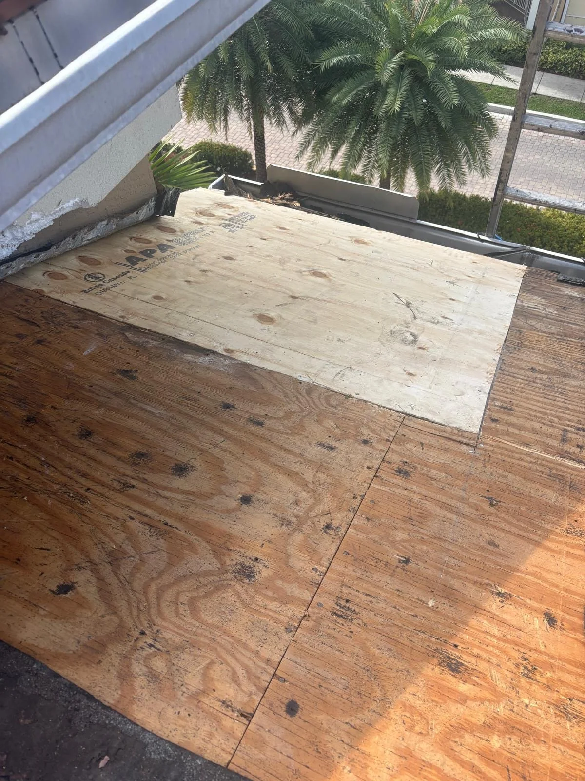 A view of a balcony or small outdoor area where wooden flooring is being installed. The image shows a transition between an existing darker wood floor and a new lighter wood floor with plywood flooring for support. In the background, there is a tree and a sidewalk outside.