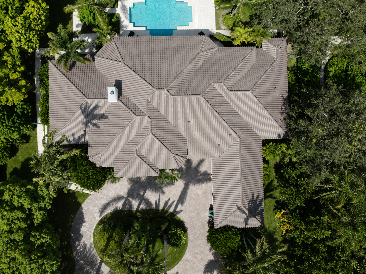 Aerial view of a house with a brown tiled roof, surrounded by lush green trees, with a paved circular driveway and a swimming pool in the backyard.