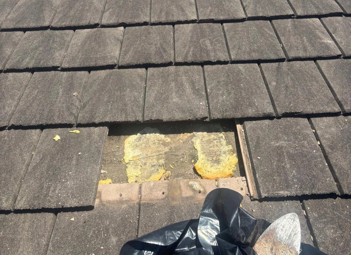 View of a roof with shingles, showing a missing section revealing insulation and underlayment underneath, with a roofing tool and trash bag nearby.
