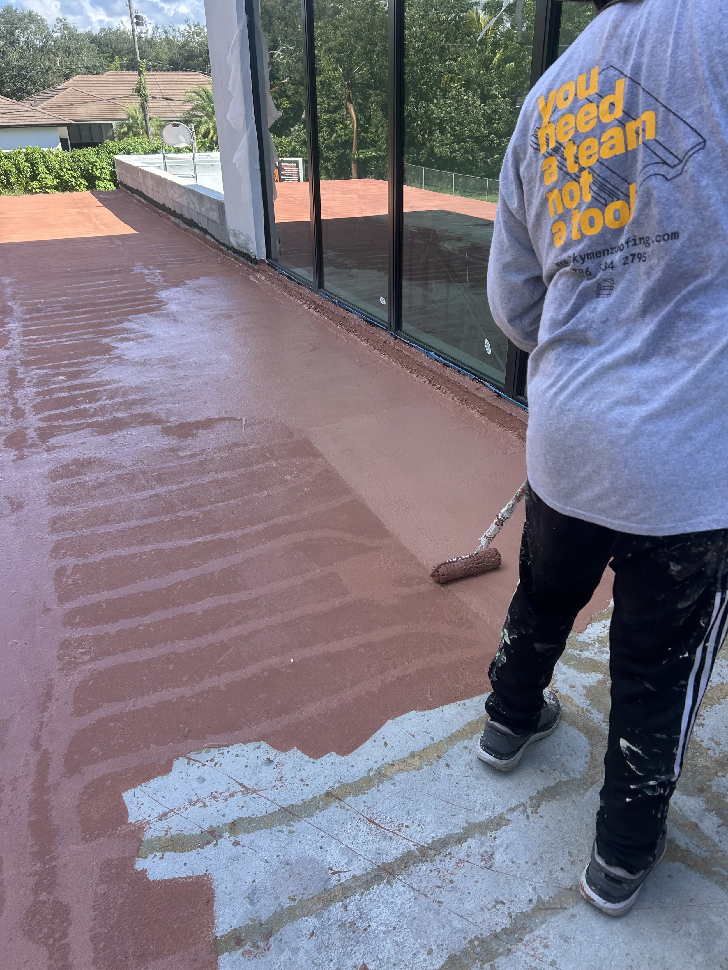 Person painting a concrete floor with a roller brush, wearing black pants and sneakers. The area appears to be a terrace or balcony with a glass railing, houses, and trees in the background.