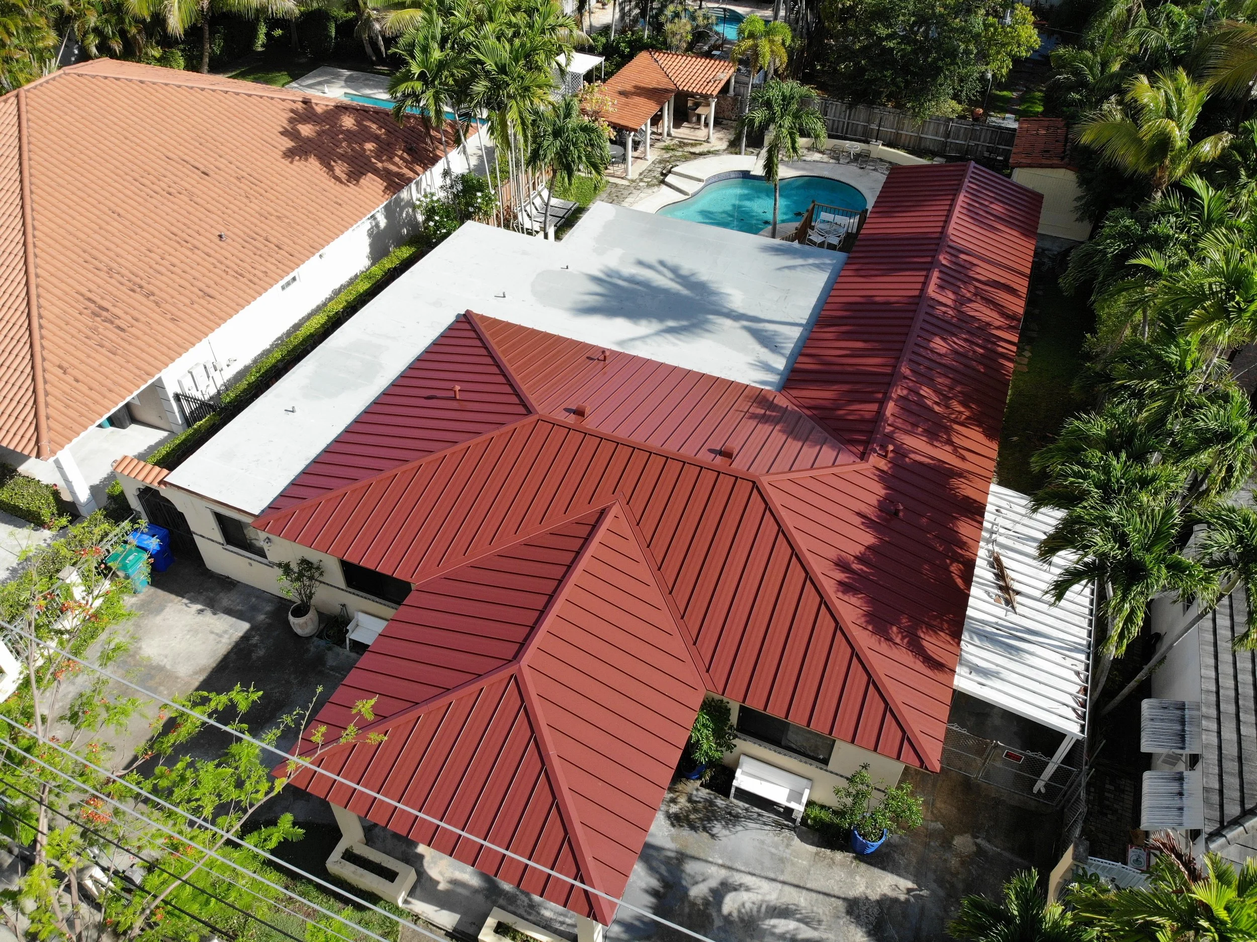 Aerial view of a residential backyard with red metal roofs, swimming pools, palm trees, and lush greenery.