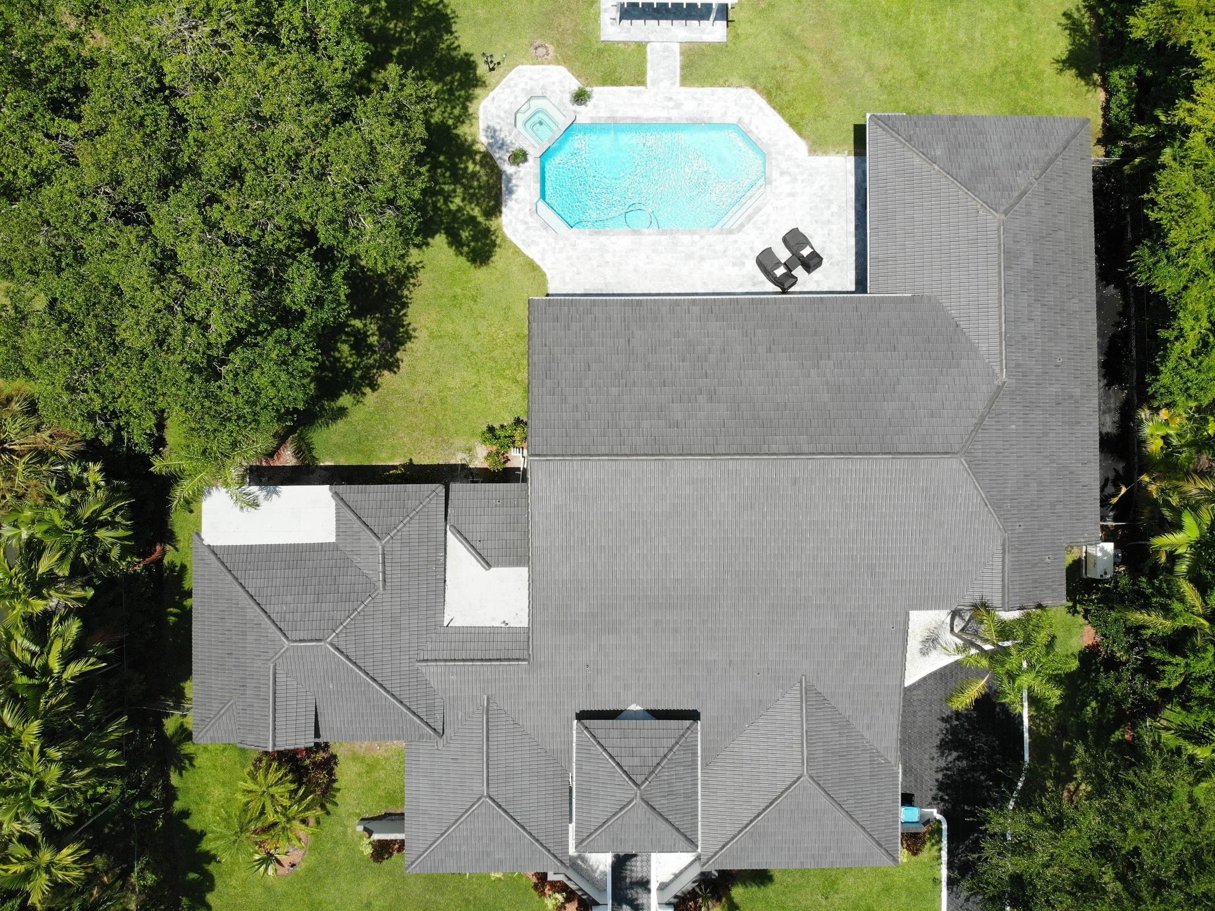 Aerial view of a house with a gray roof, a backyard with a swimming pool, a hot tub, and surrounding lush green trees and grass.