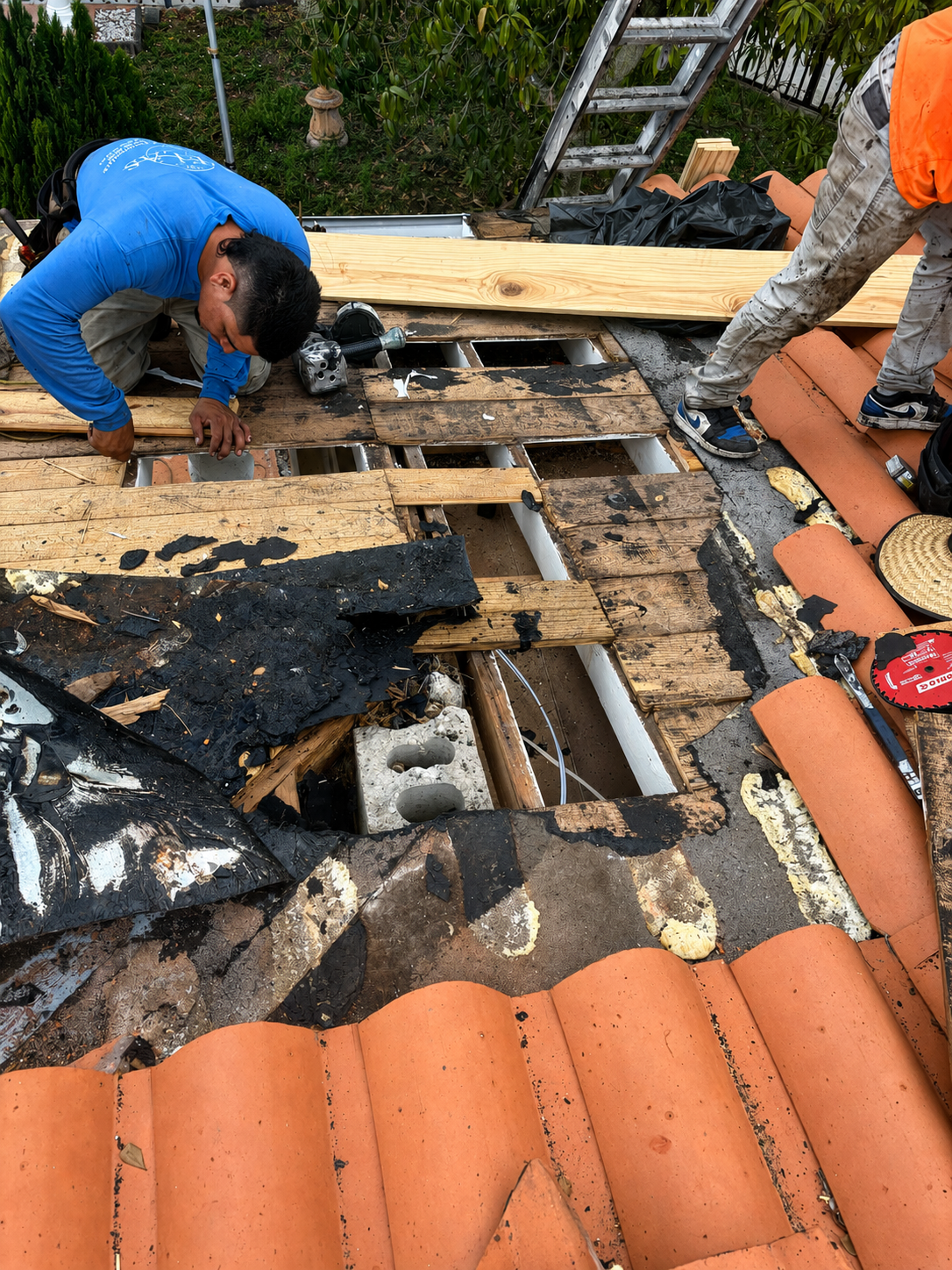 Two workers repairing a roof, exposing wooden beams and underlayment, with building supplies and tools around them.