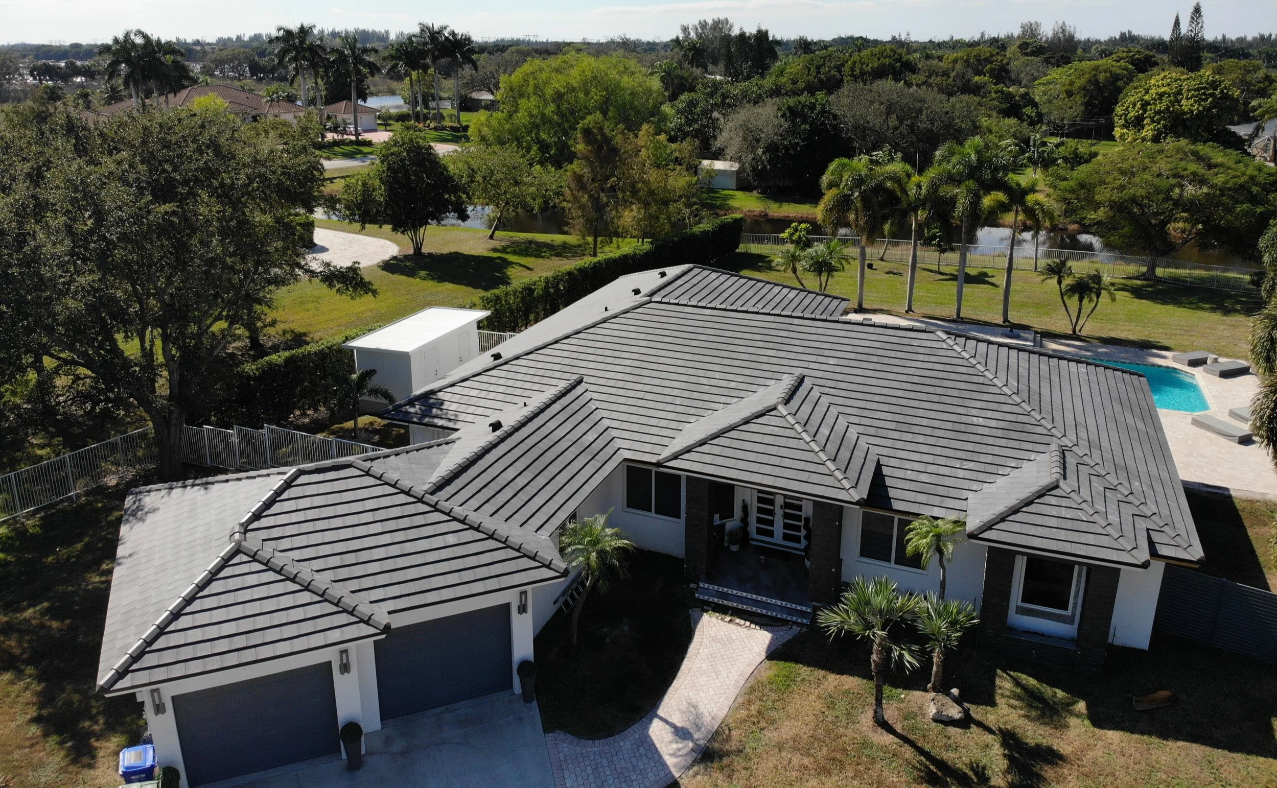 Aerial view of a modern house with gray roof, surrounded by green trees, a swimming pool, and a fenced backyard in a lush neighborhood.