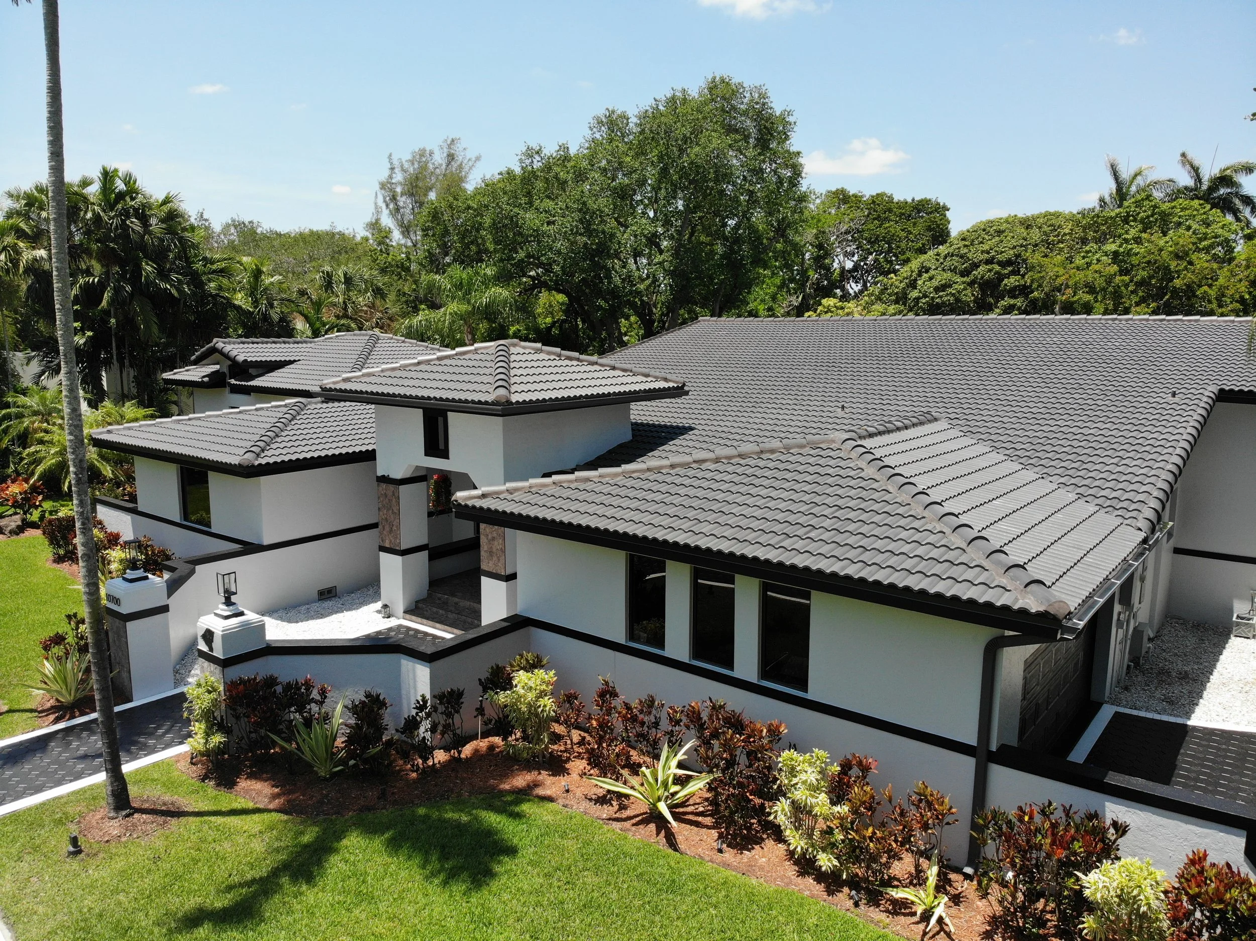 A modern house with a gray tiled roof, white walls, and large windows, surrounded by a well-maintained green lawn and various shrubs and trees, under a clear blue sky.