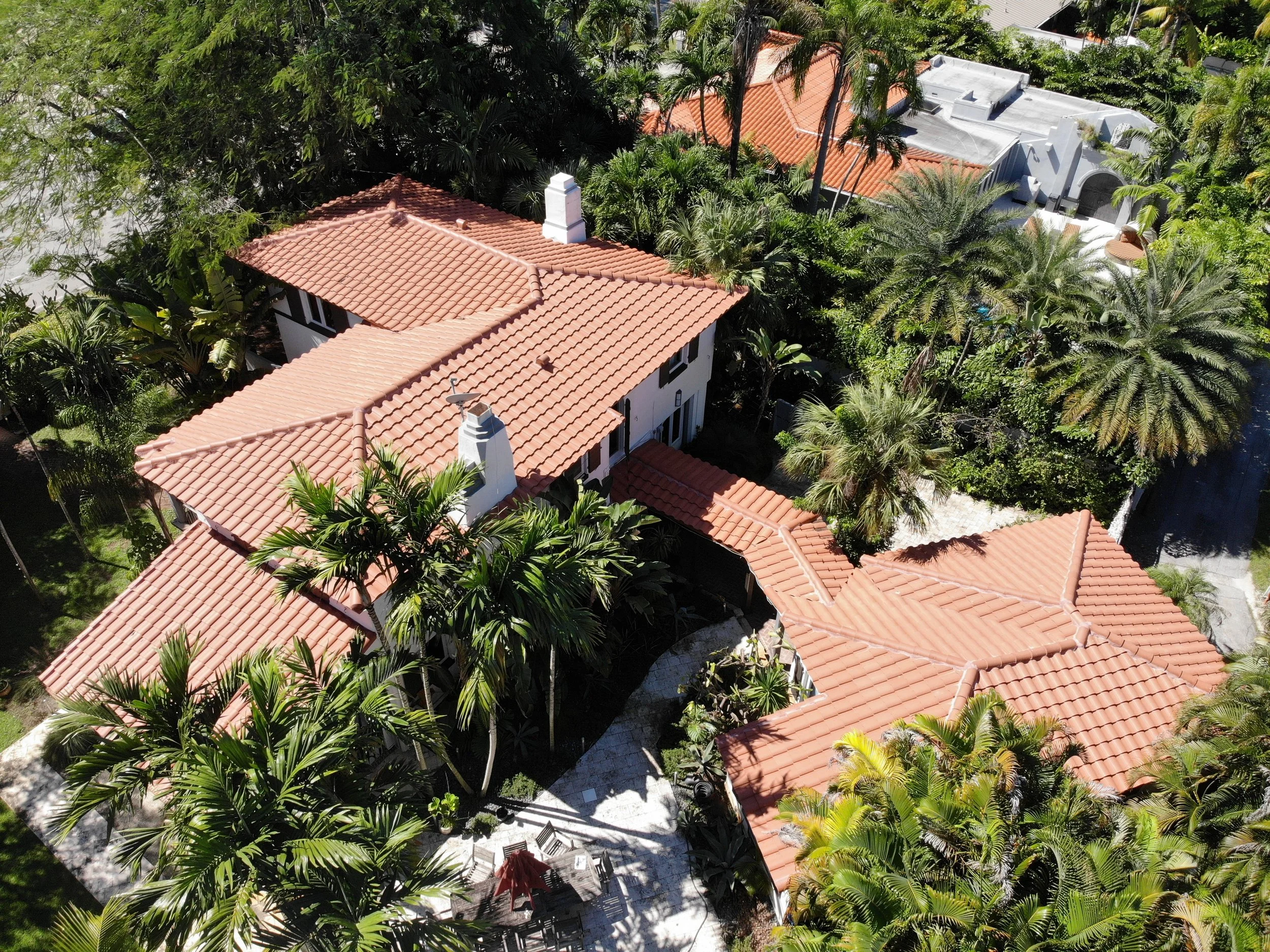 Aerial view of a lush, green residential area with houses featuring red tile roofs, surrounded by tall palm trees and other tropical vegetation.