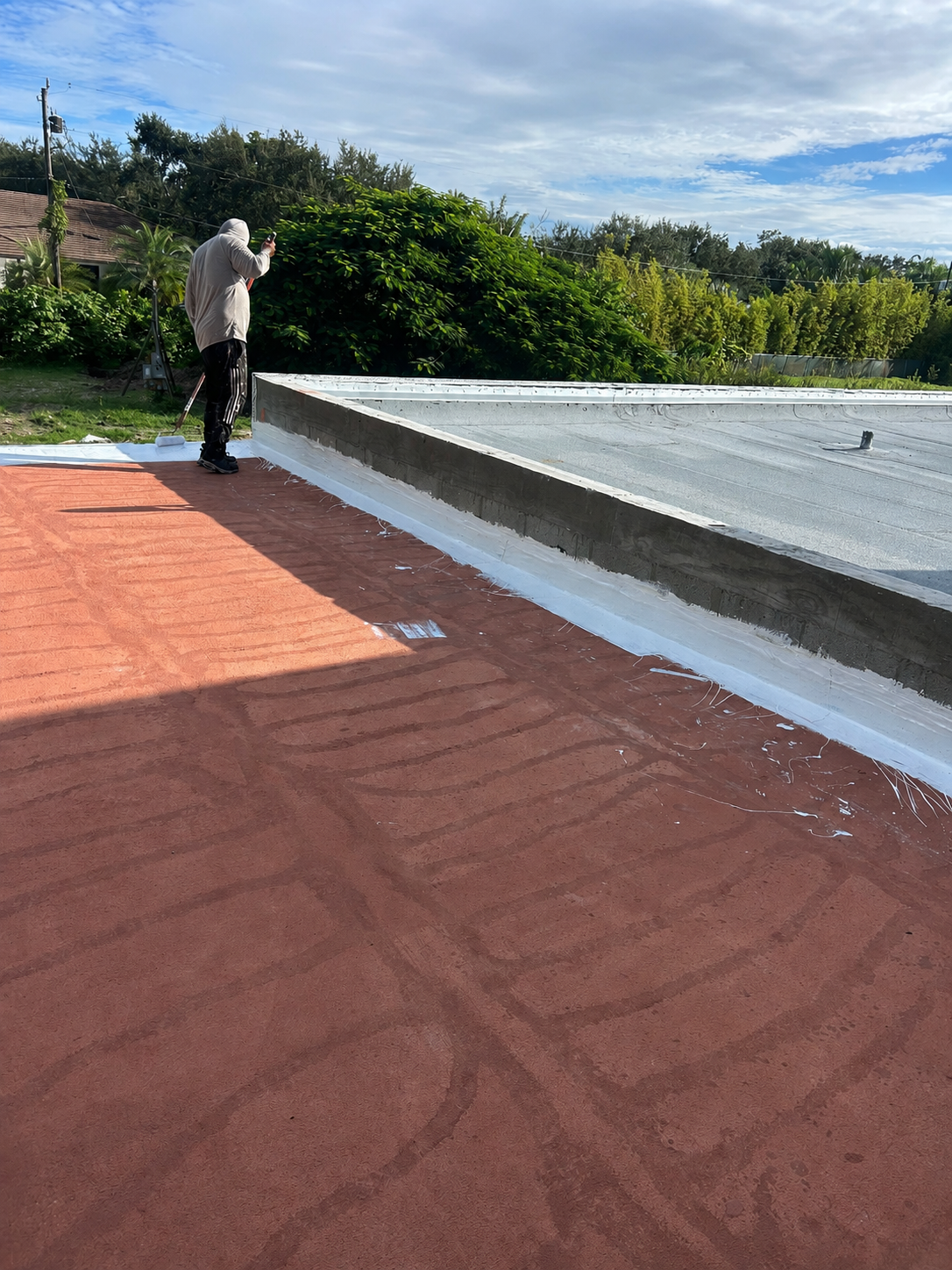 A person in a hoodie and dark pants is painting or sealing the edge of a flat roof with a white coating, while standing on a reddish-brown surface with visible tire tracks or patterns, under a partly cloudy sky, with green trees and bushes in the background.