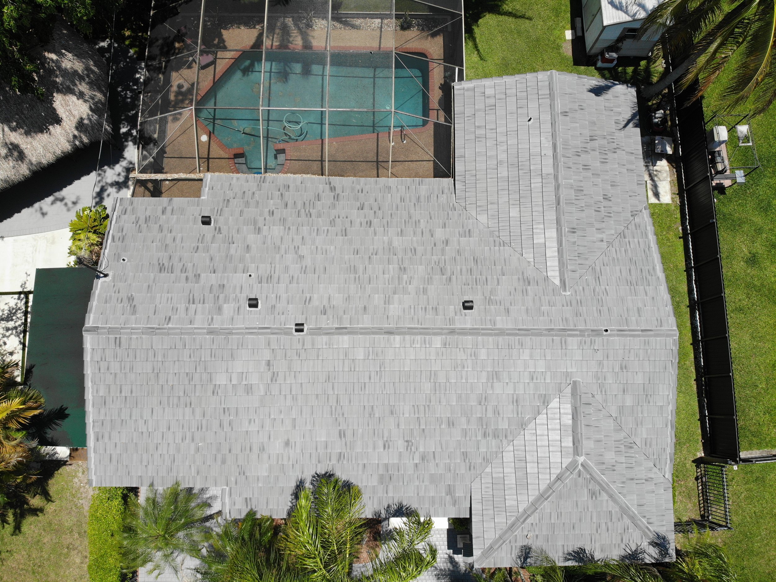 A house with a grey tiled roof and a backyard enclosed by a black fence, featuring a swimming pool with a screened enclosure, surrounded by grassy yard with trees and outdoor furniture visible.