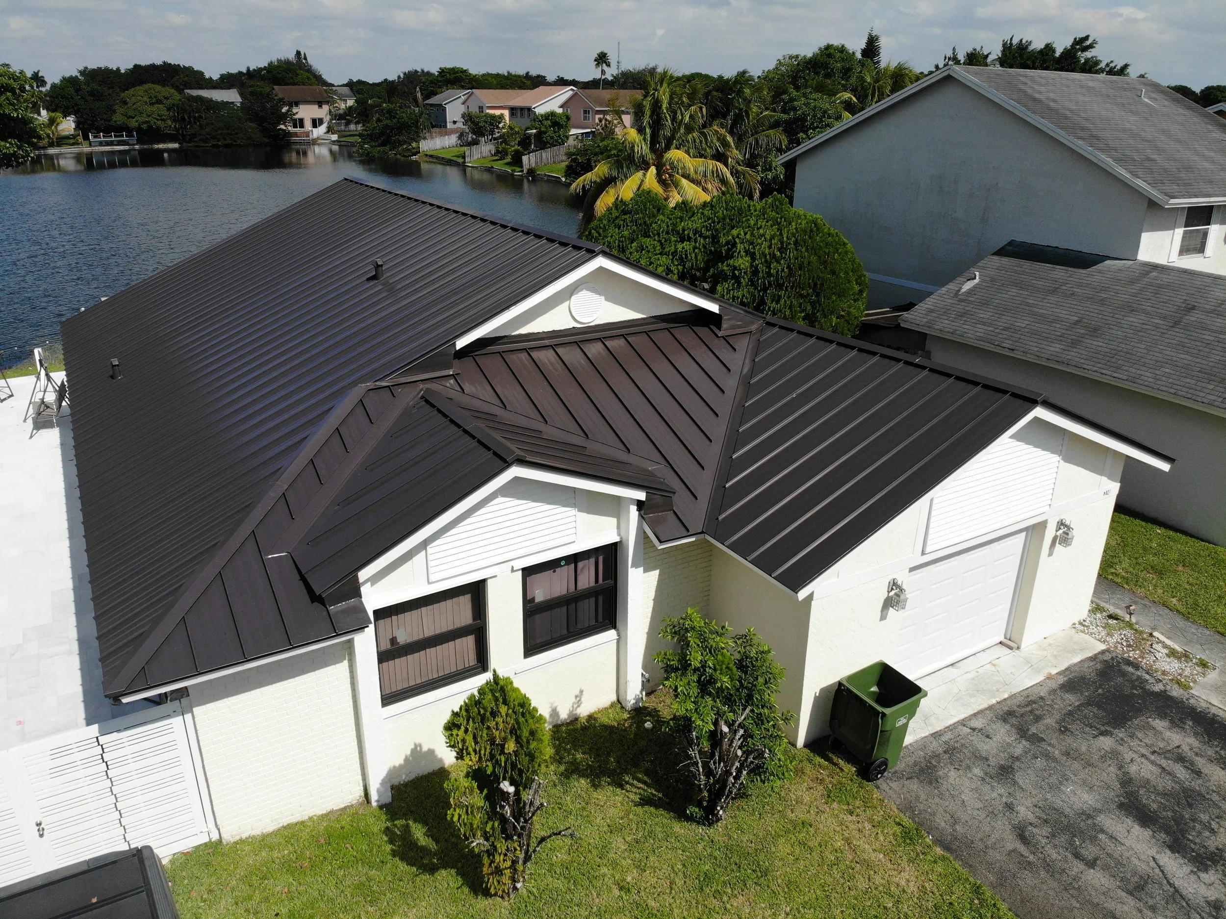 Aerial view of a residential neighborhood with houses, a pond, trees, and a driveway with a trash bin.