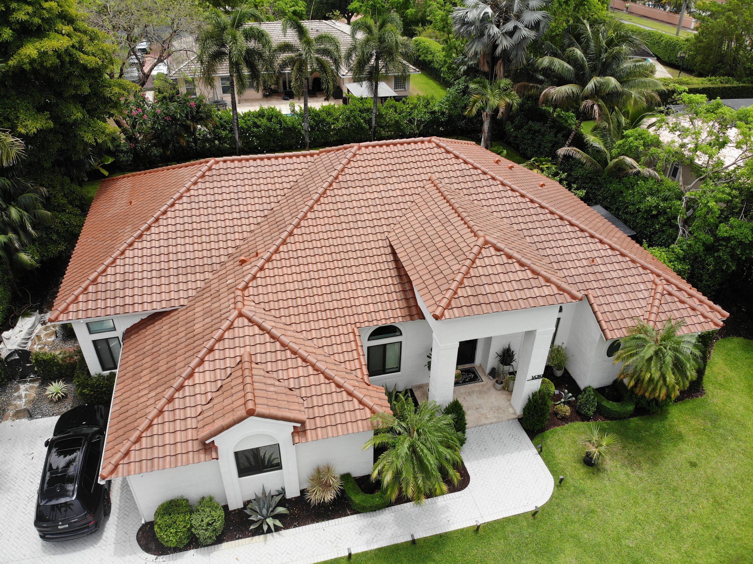 Aerial view of a house with a red-tile roof, surrounded by lush green trees and plants. The house has a white exterior with large windows and a driveway with a parked black car.
