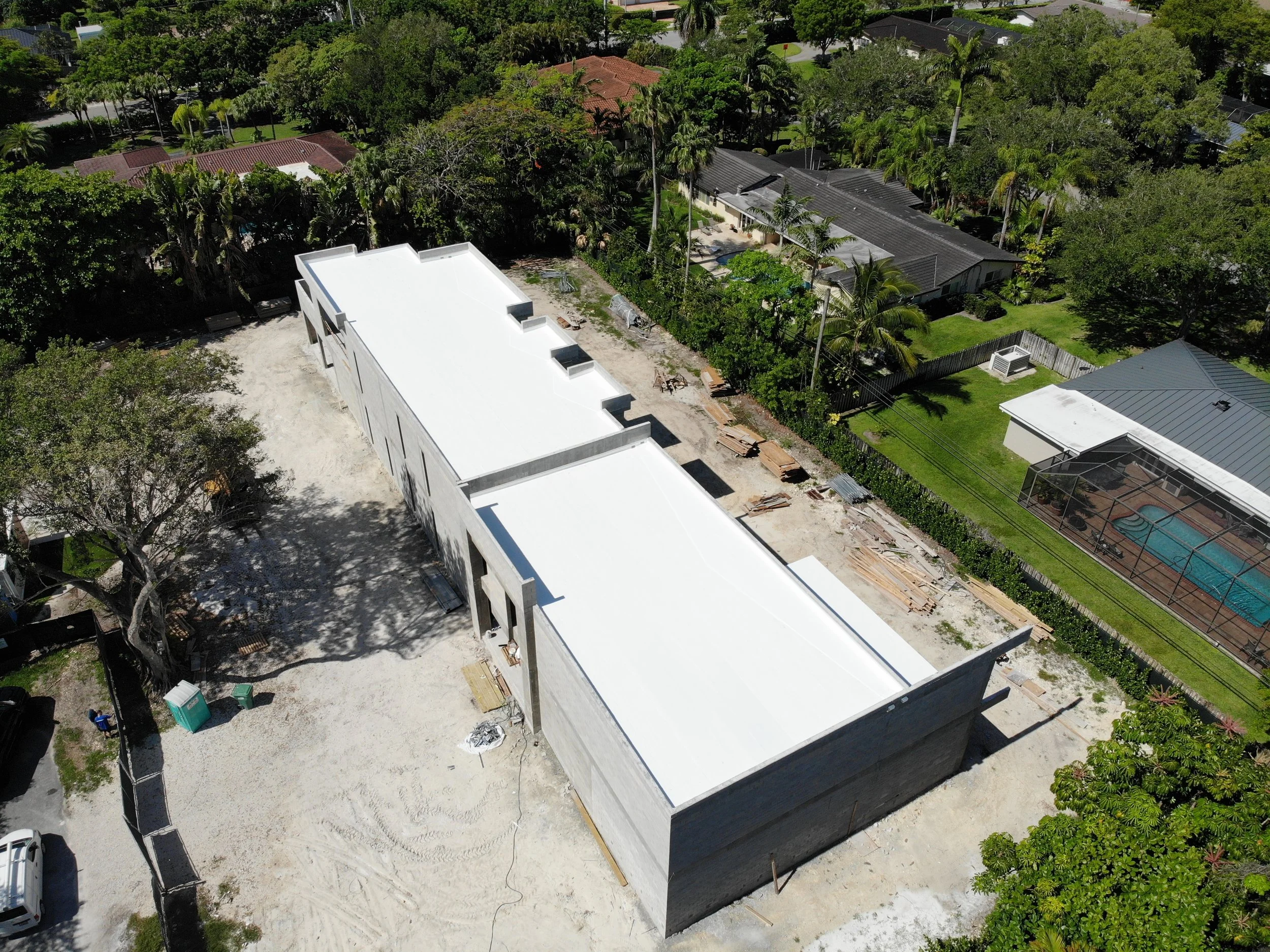 An aerial view of a construction site with white building structures, surrounded by trees and neighboring houses with swimming pools.