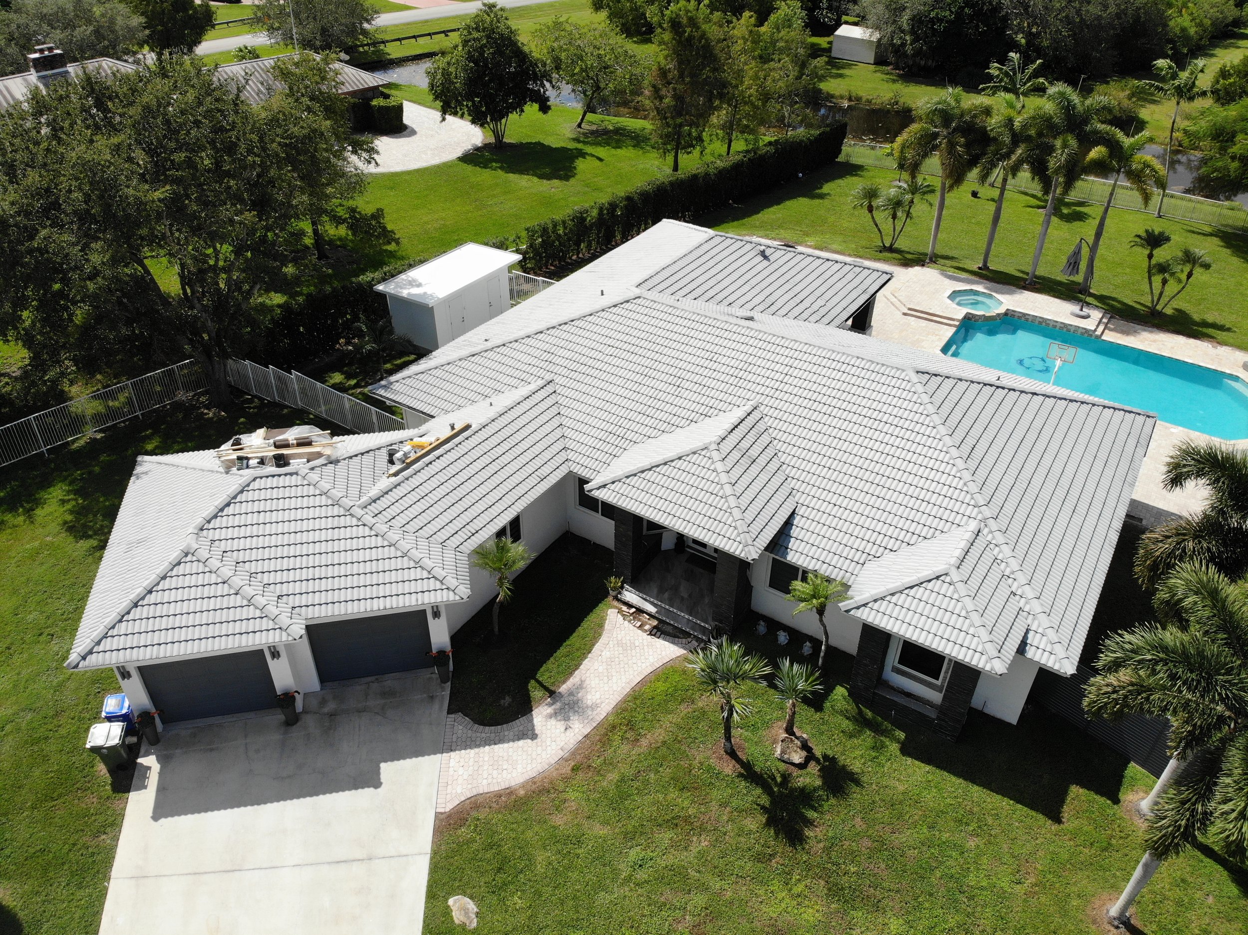 An aerial view of a residential backyard with a white house, swimming pool, lush green lawn, palm trees, and surrounding landscape.