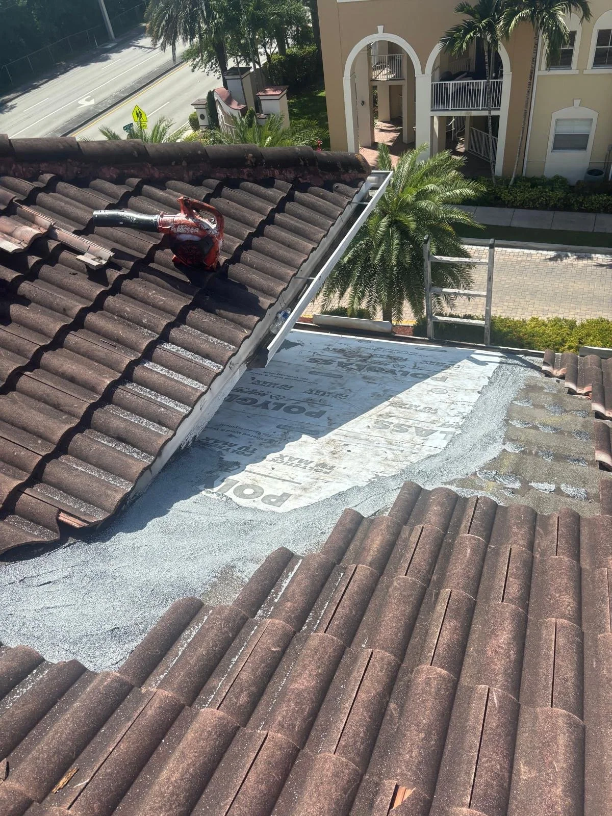 View from a rooftop showing weathered clay roof tiles, a section under repair with new roofing material, a vent pipe, palm trees, a residential street, and an apartment building in the background.