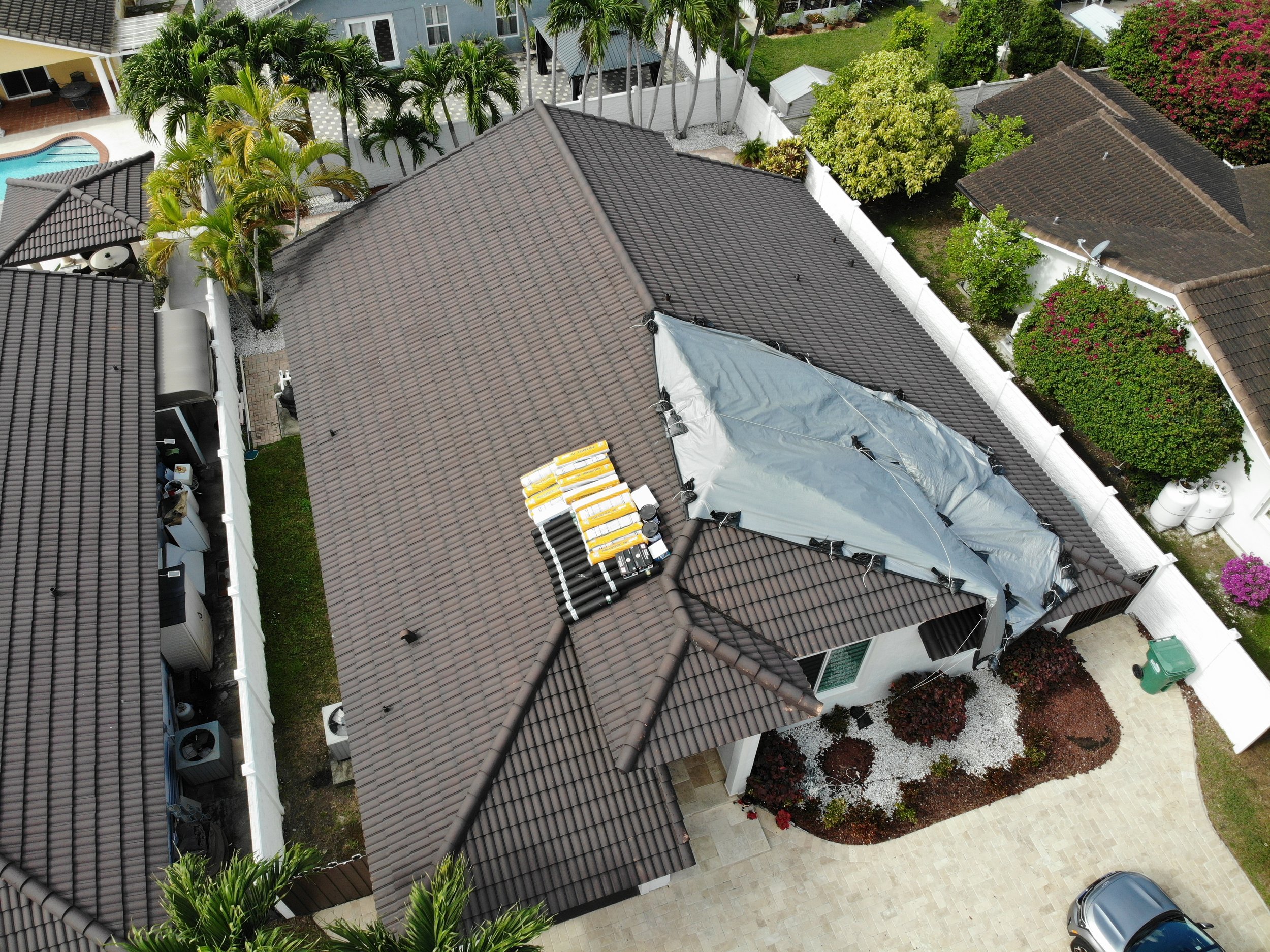 An aerial view of a backyard with a house, a swimming pool, palm trees, and garden areas with colorful plants and trees.