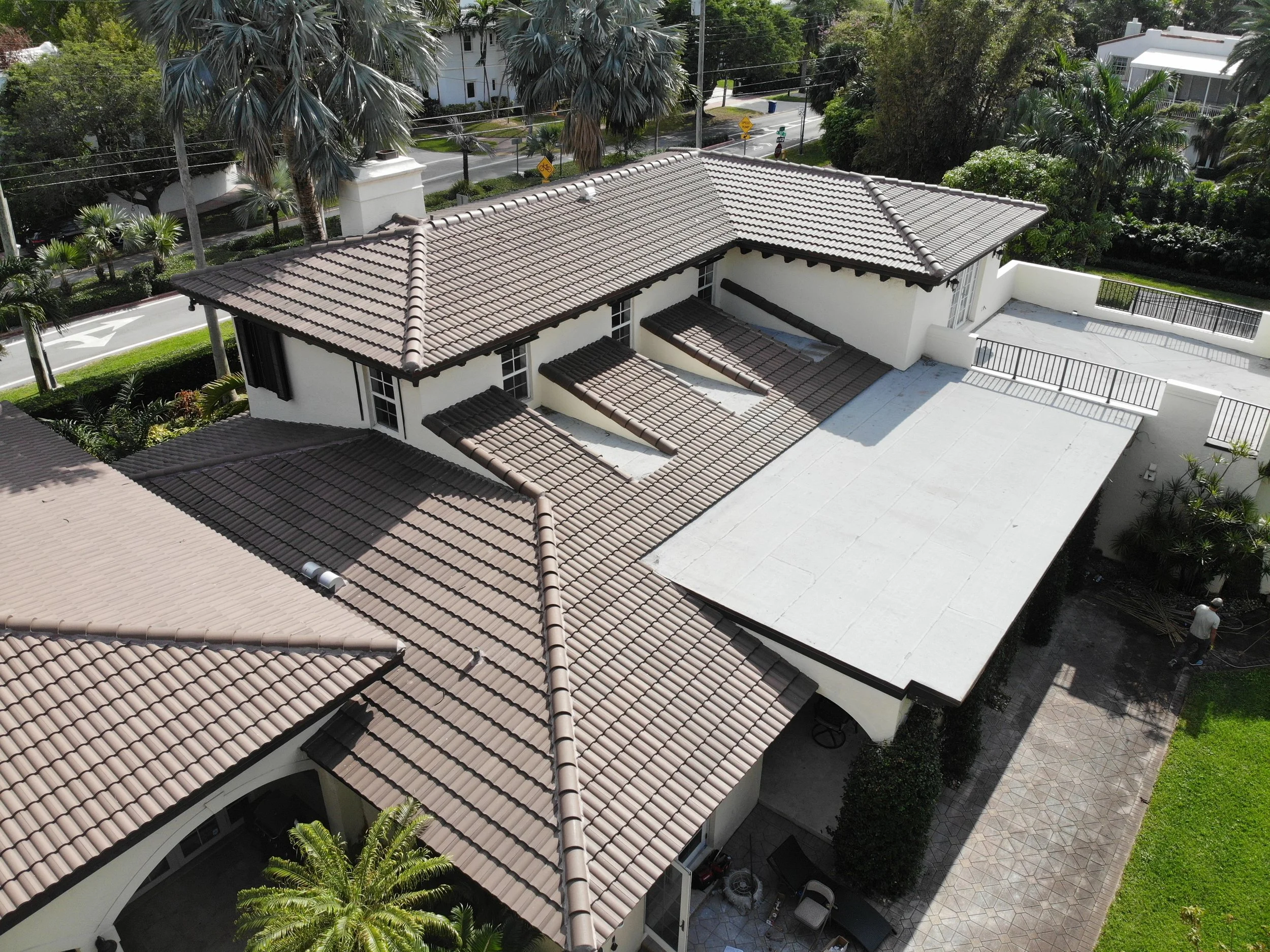 Aerial view of a house with a terracotta tiled roof, a white patio, surrounding tropical trees, and a street with bike lane nearby.