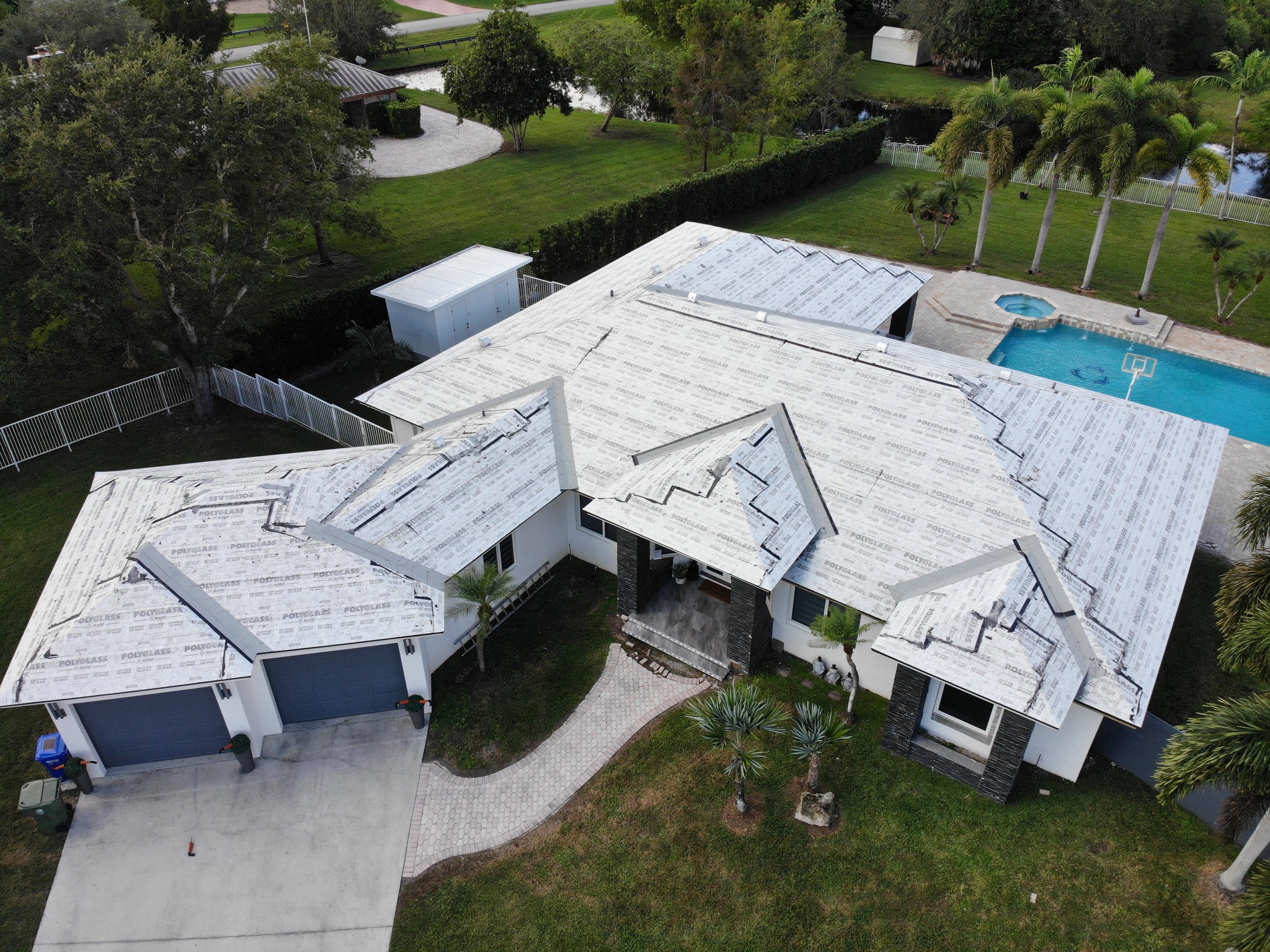 An aerial view of a house under construction with roofing materials, a swimming pool, palm trees, and green landscaping.