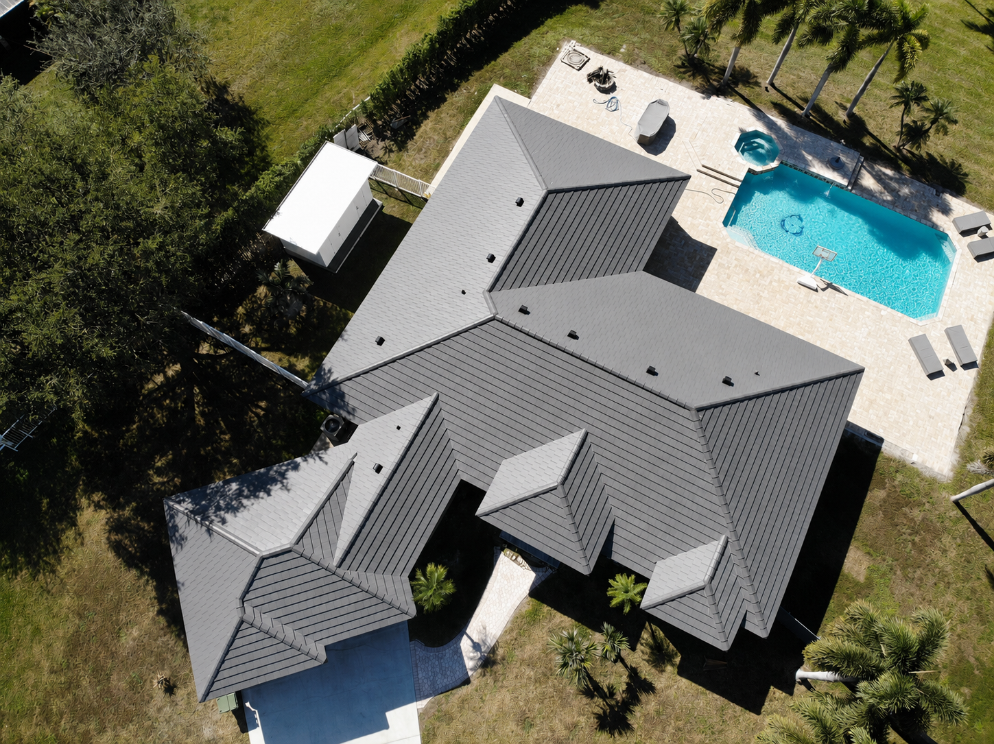 Aerial view of a house with gray metal roof, surrounded by trees, with a backyard featuring a swimming pool, lounge chairs, and a hot tub.