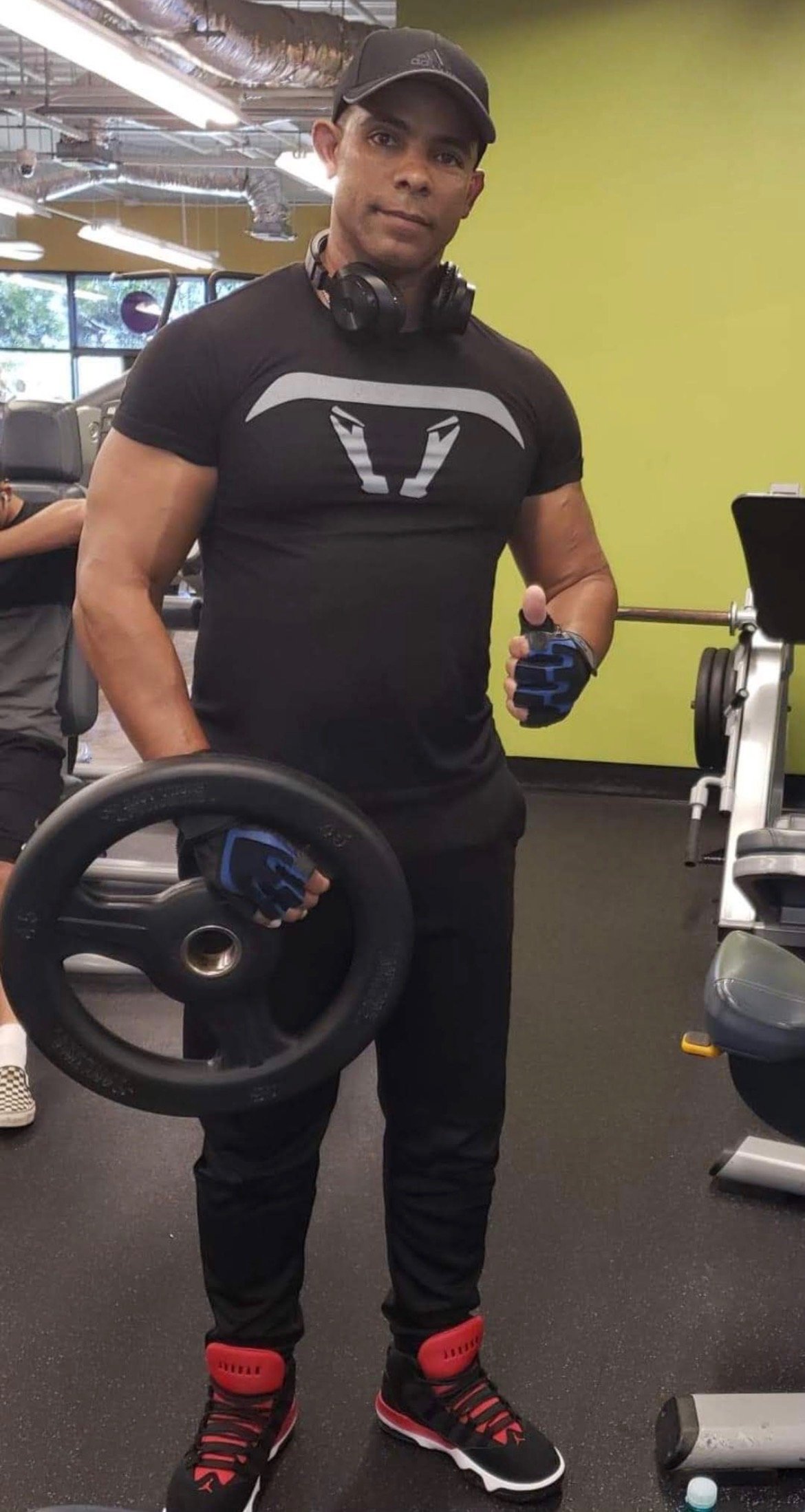 A man at the gym holding a weight plate and wearing workout clothes, including a cap, headphones, and sneakers, with gym equipment and windows in the background.