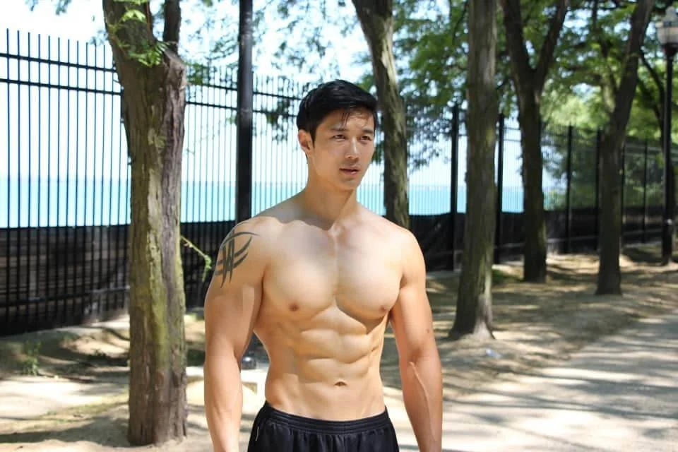 A shirtless man with defined abs and a tribal tattoo on his left shoulder, standing outdoors among trees with a fence in the background.