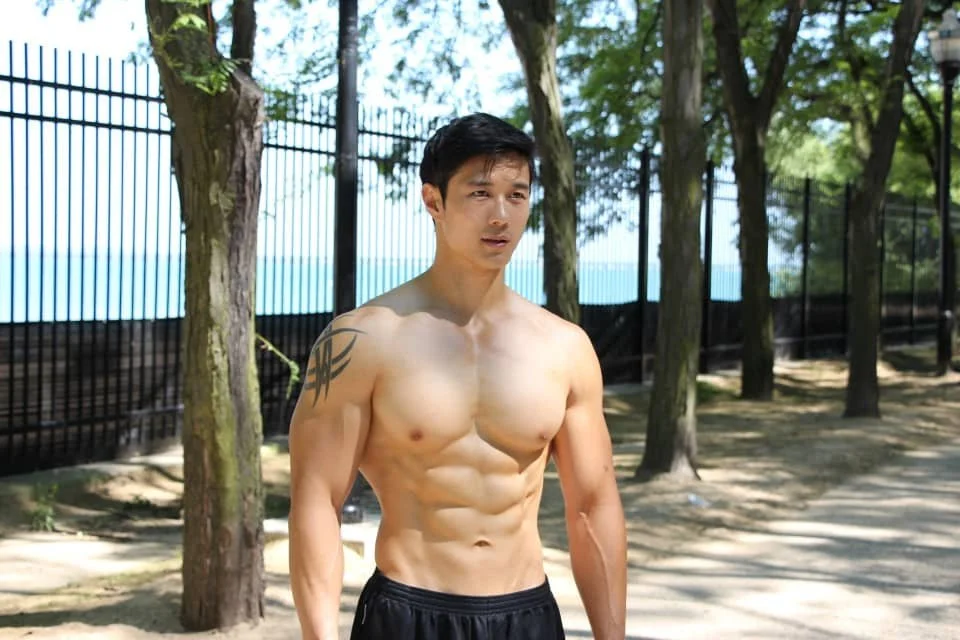 A shirtless man with black hair standing outdoors near trees, with a fence in the background.