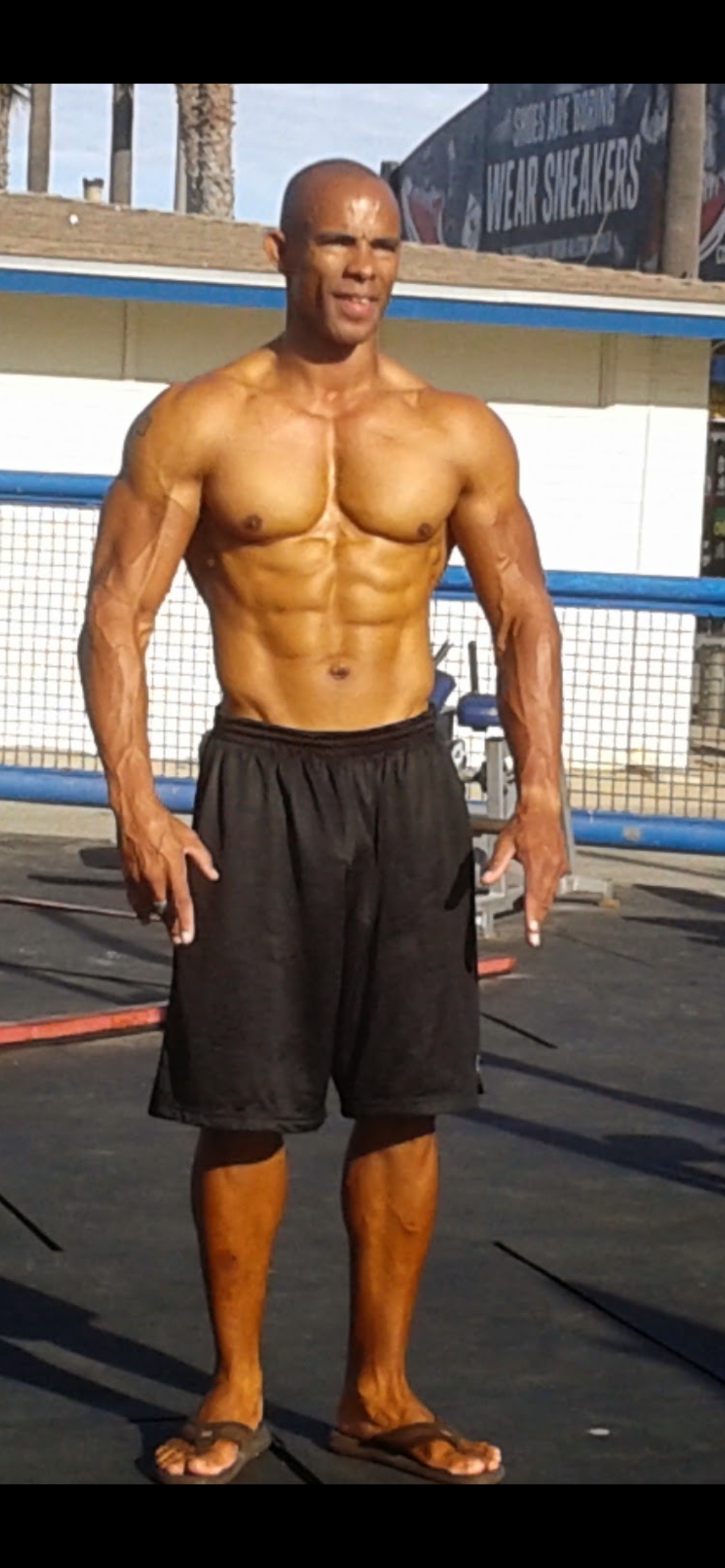 A muscular, shirtless man with a bald head and dark skin standing outdoors in shorts and flip-flops, with fencing and a sign for Nike sneakers in the background.