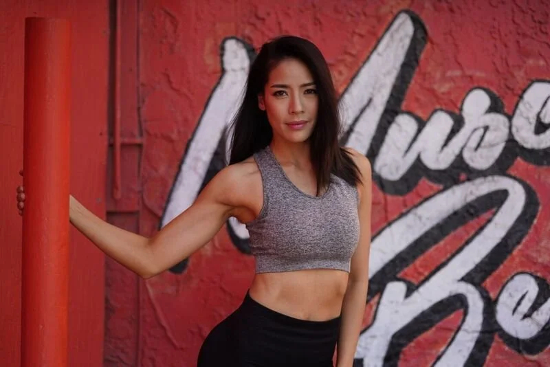 A woman in a gray crop top and black leggings standing outdoors against a red wall with white graffiti.