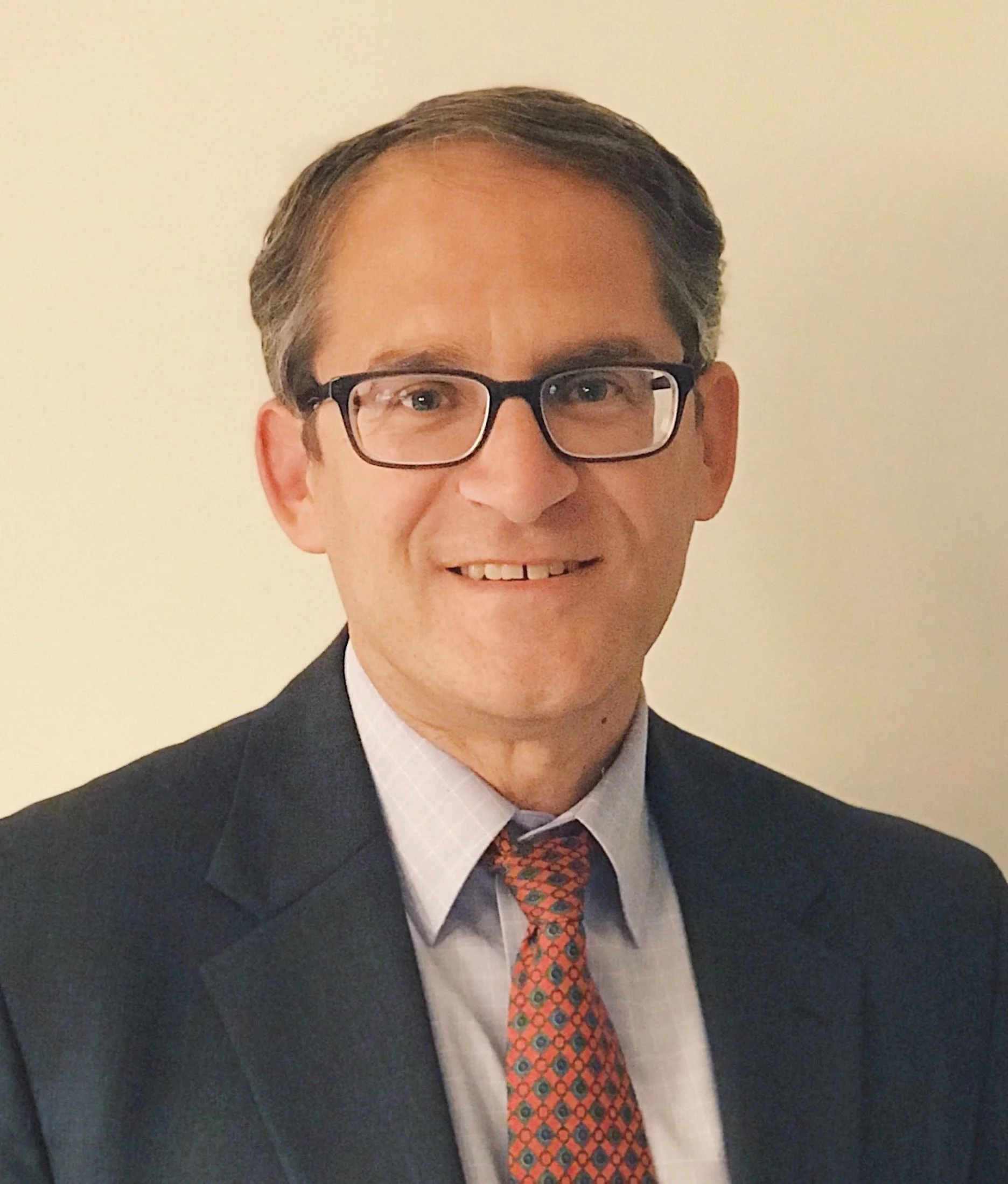 A man wearing glasses, in formal attire with a dark suit, white shirt, and a colorful patterned tie, smiling in front of a plain light-colored background.