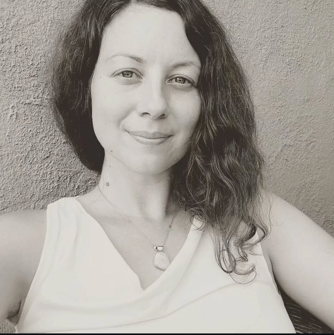 A woman with wavy hair and a necklace, smiling softly, taking a selfie against a textured wall.
