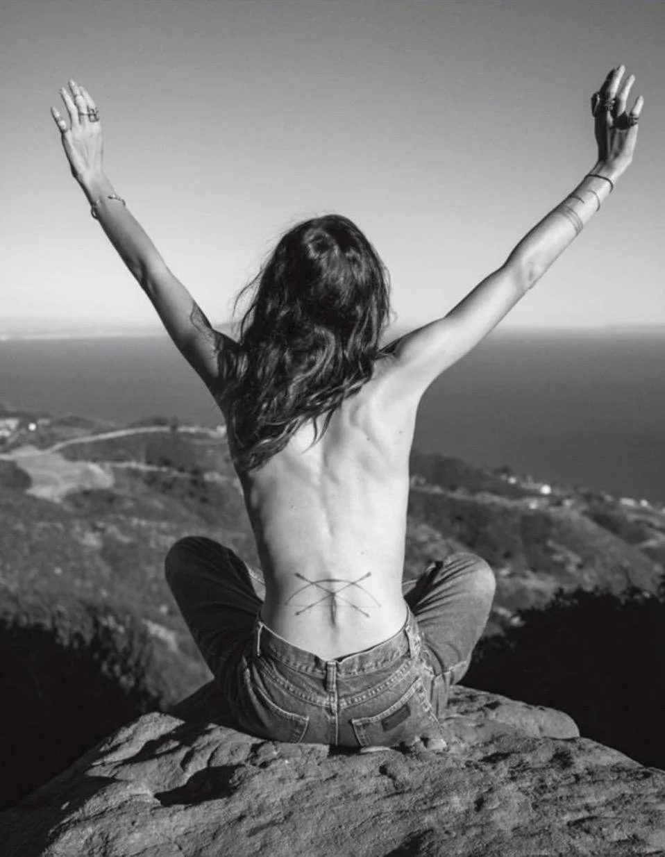 A woman with long hair sitting on a rock with her back facing the camera and arms raised, overlooking a coastal landscape in black and white.